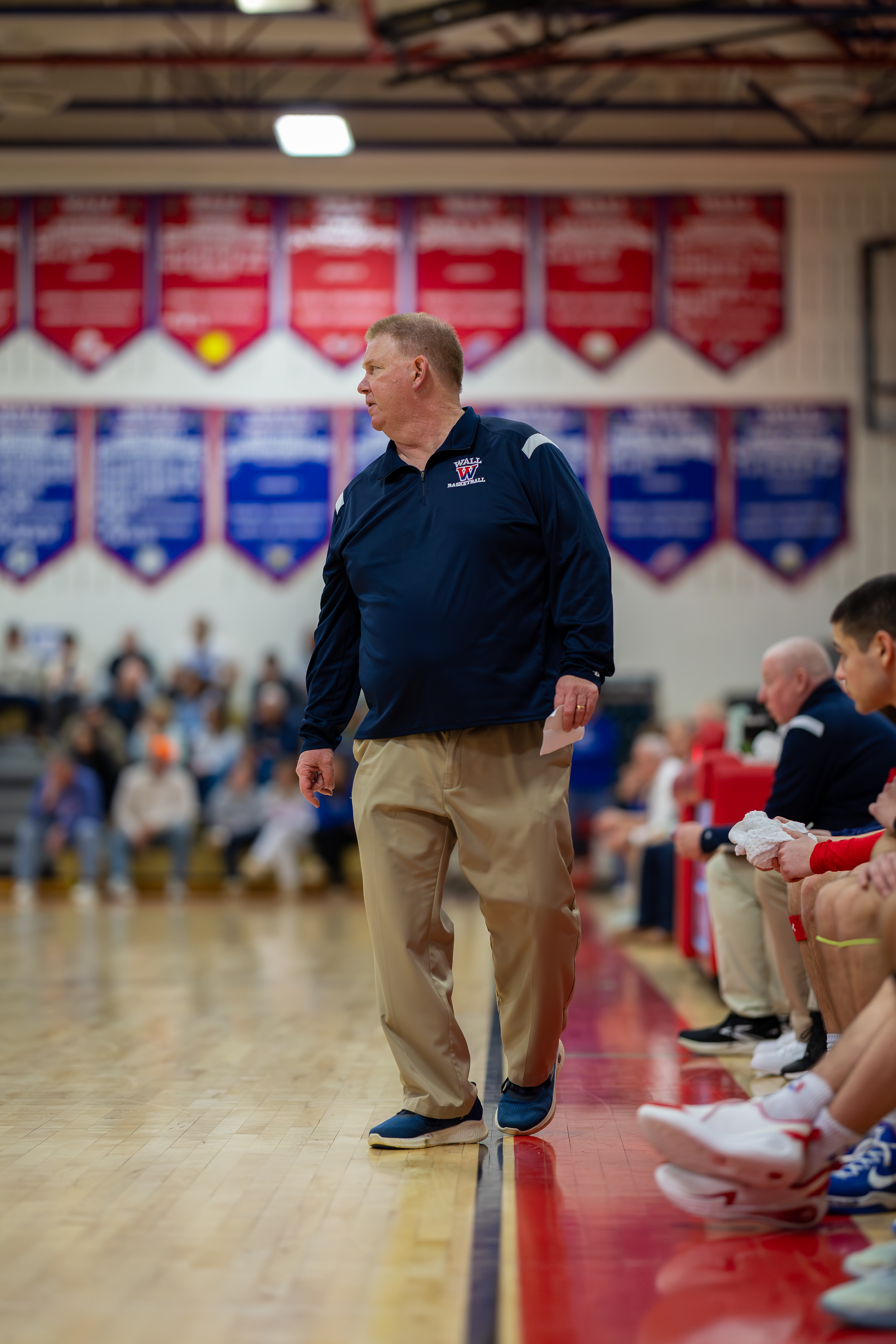 A basketball coach in a navy shirt and khaki pants stands on the court, while spectators watch from the bleachers in the background.