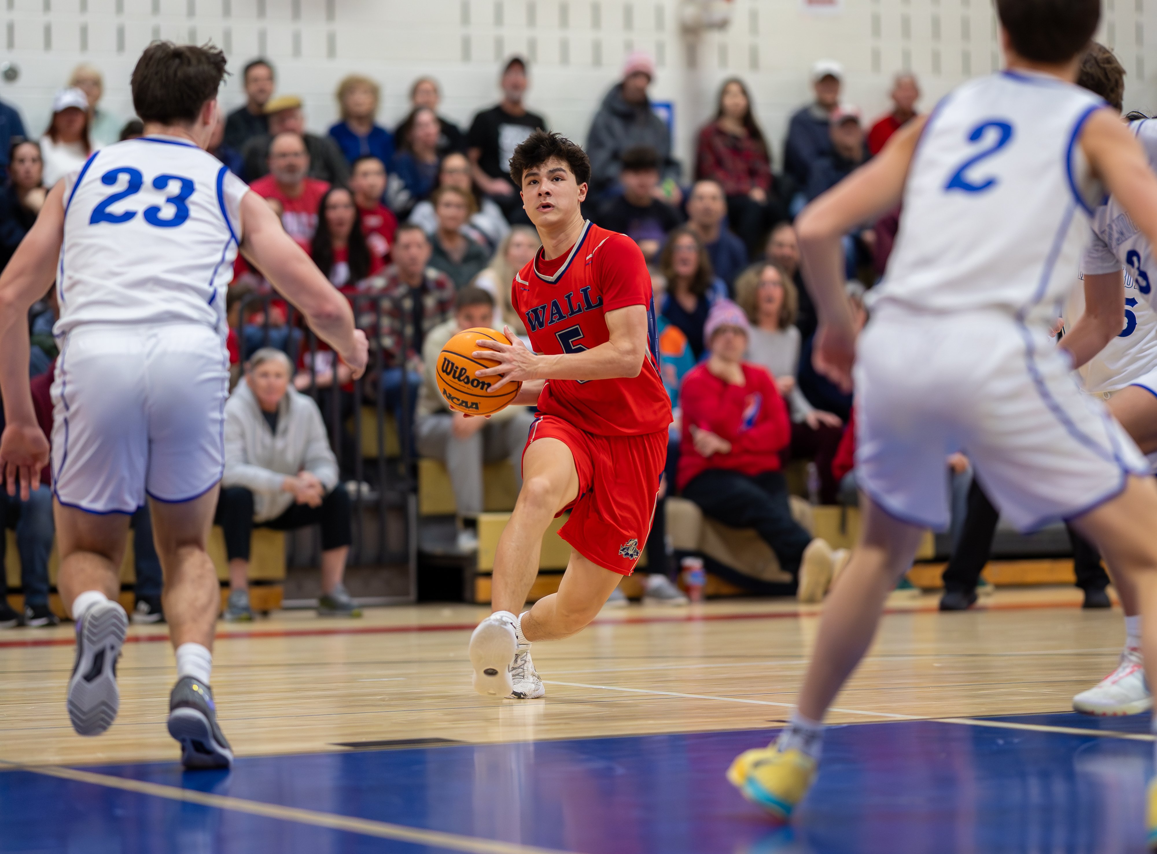 A basketball player in a red jersey dribbles the ball on a court with spectators in the background.