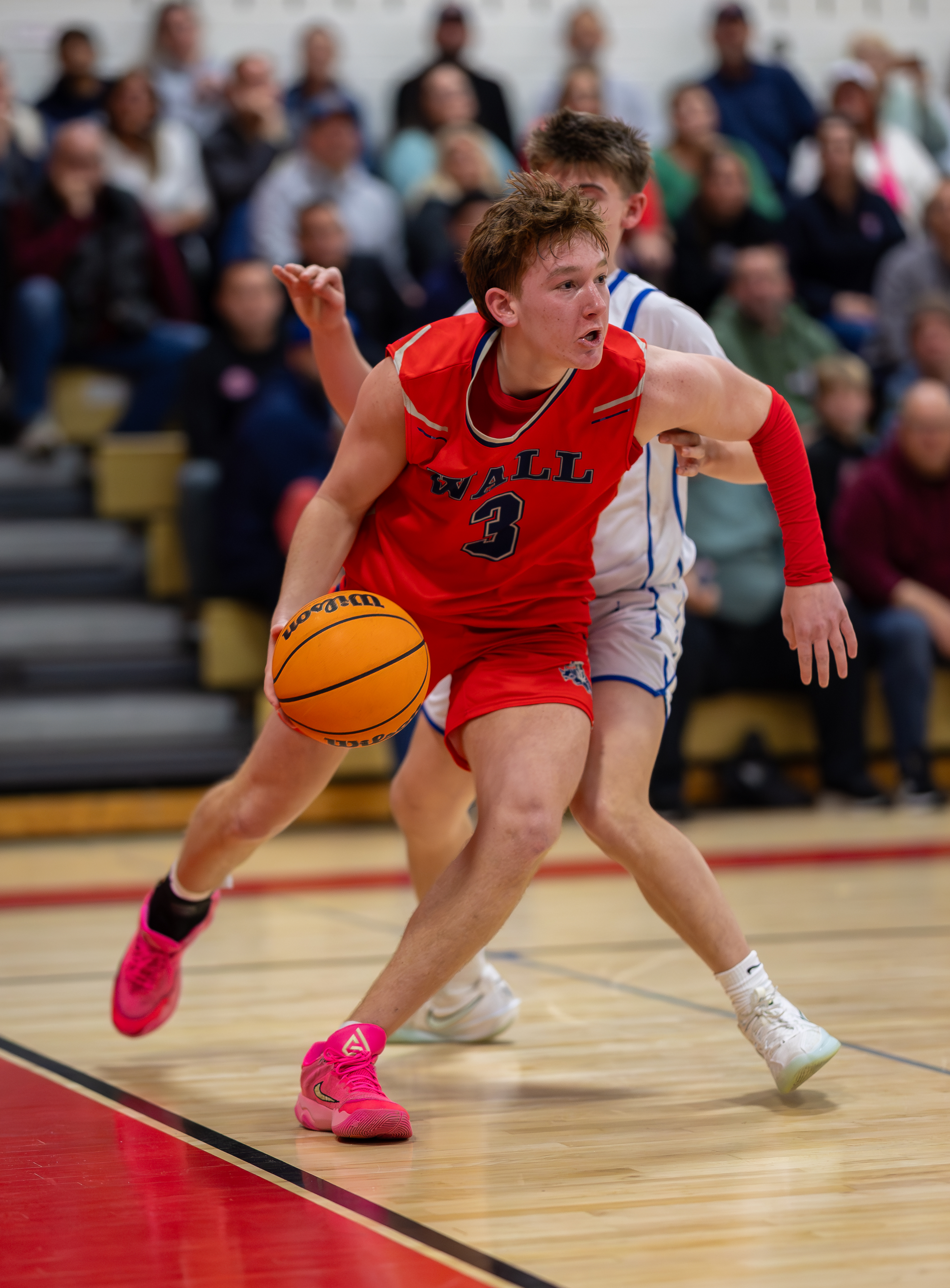 A young basketball player in a red jersey dribbles the ball on a wooden court while spectators watch from the bleachers in the background.