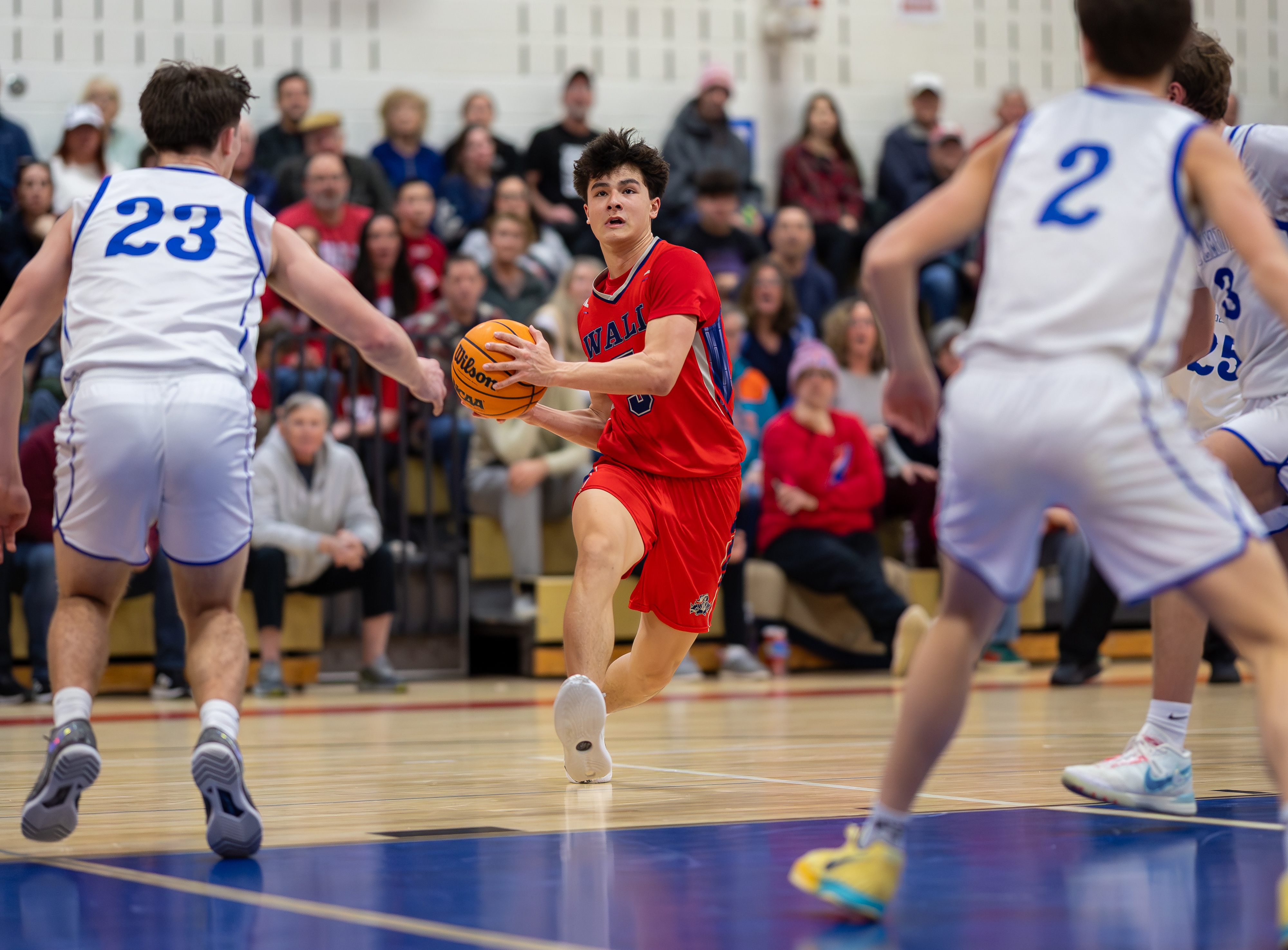A young basketball player in a red jersey dribbles the ball on a wooden court, with spectators watching from bleachers in the background.
