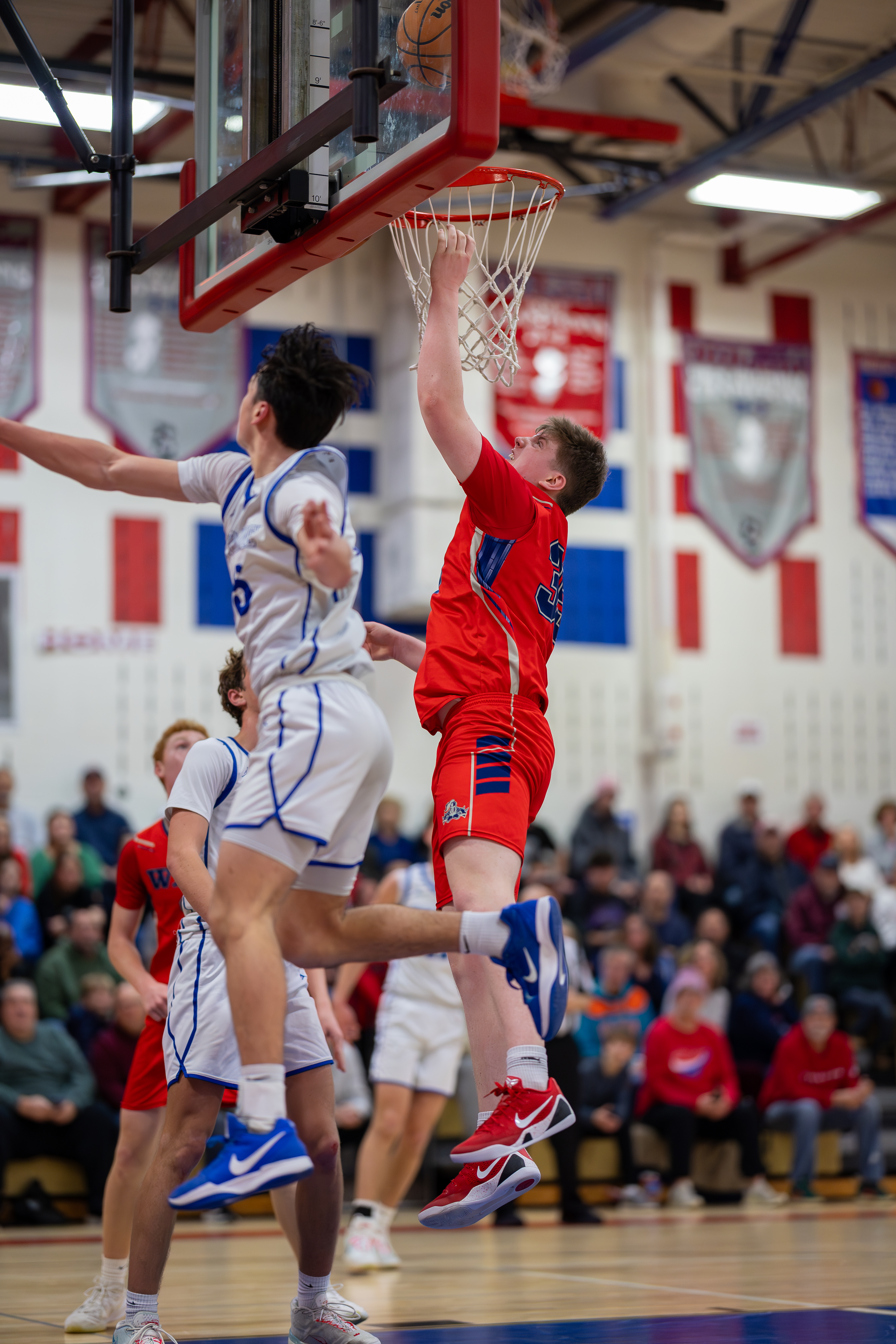 Two basketball players in red and white uniforms jump to score a basket in a crowded gymnasium.