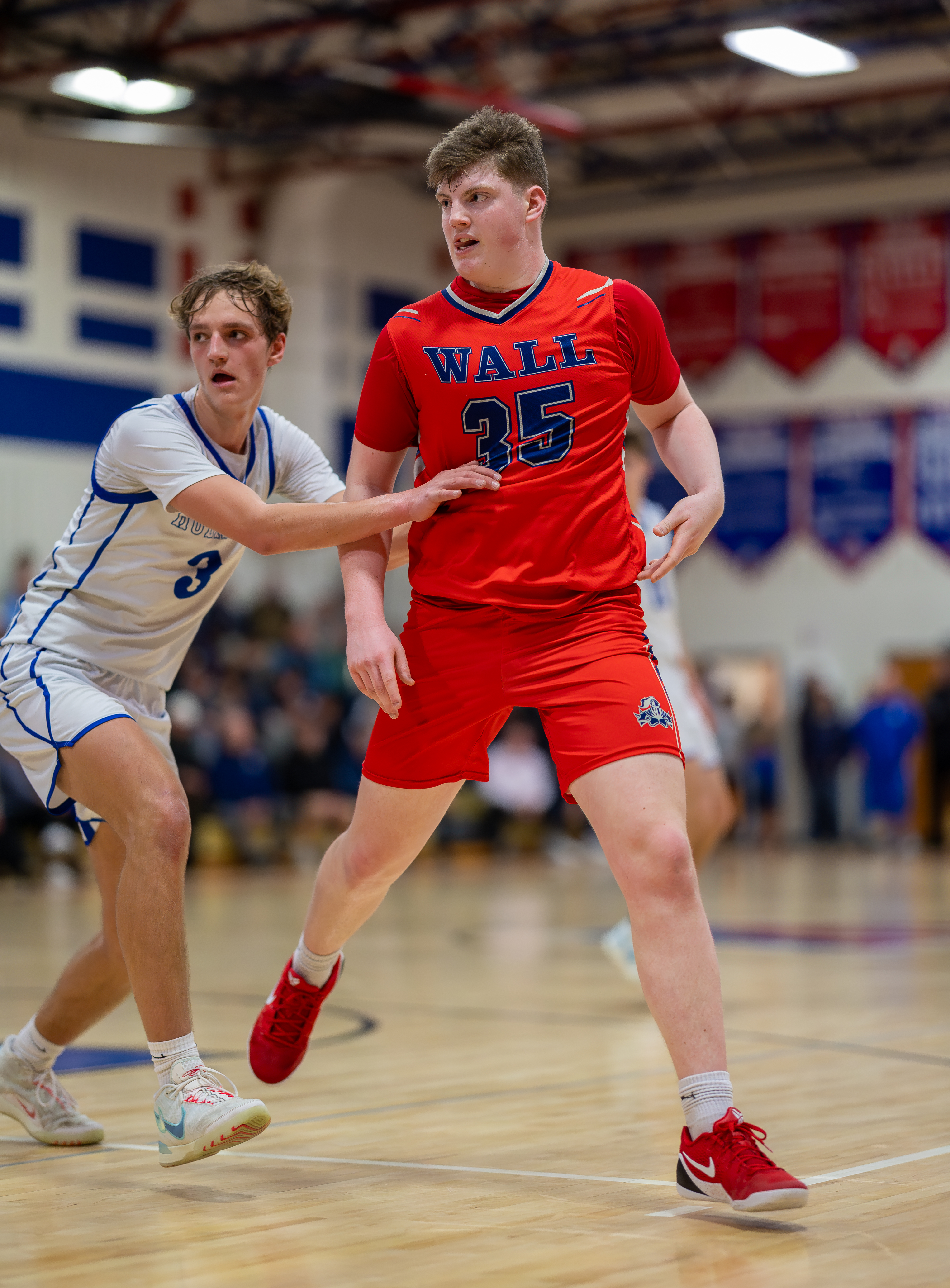 Two young basketball players, one in a red jersey with "WALL 35" on it and the other in a white jersey, are on a wooden court with blurred spectators in the background.