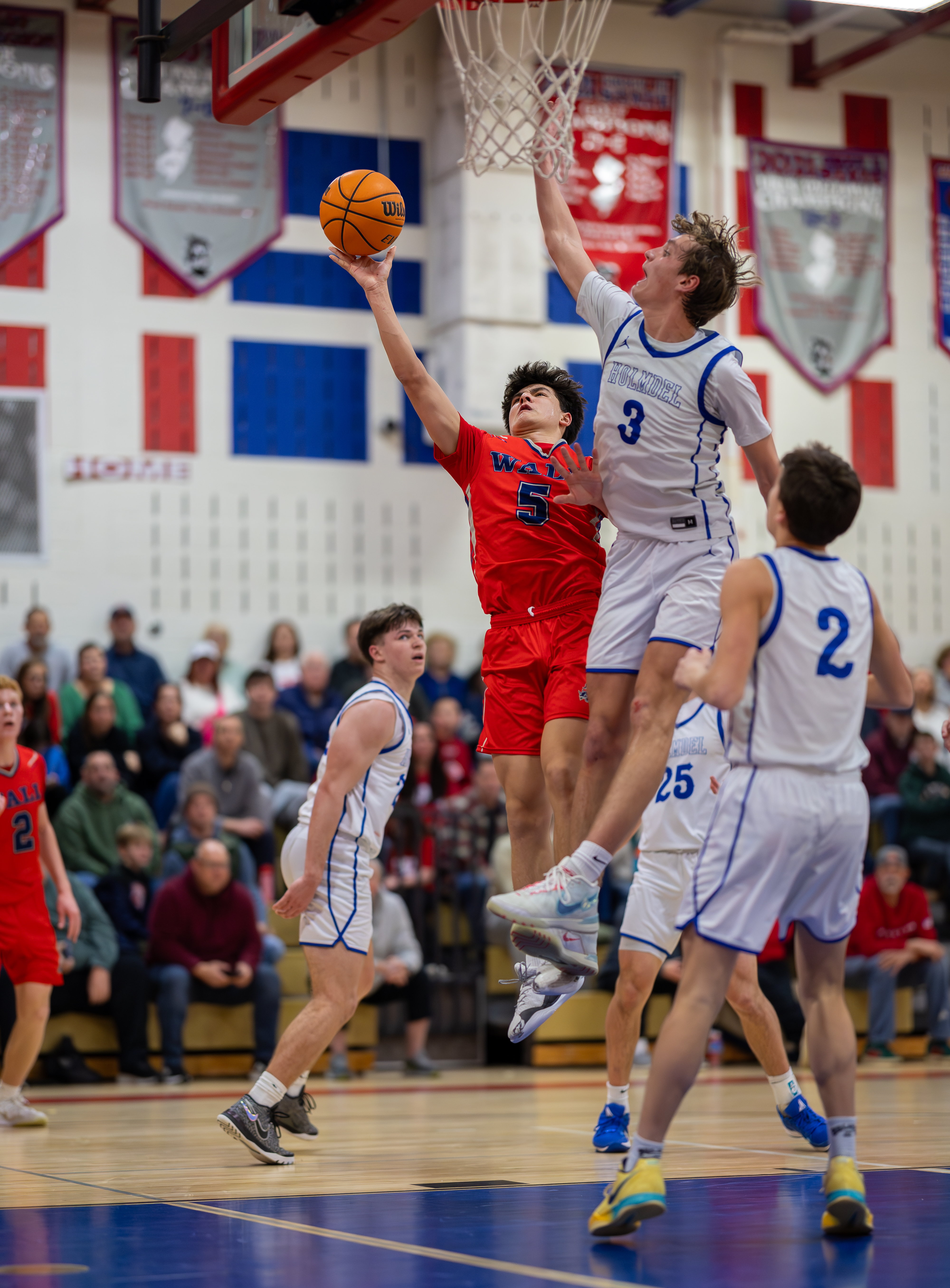 In the foreground, basketball players in red and white uniforms leap for a shot, while the background shows spectators in a gymnasium.