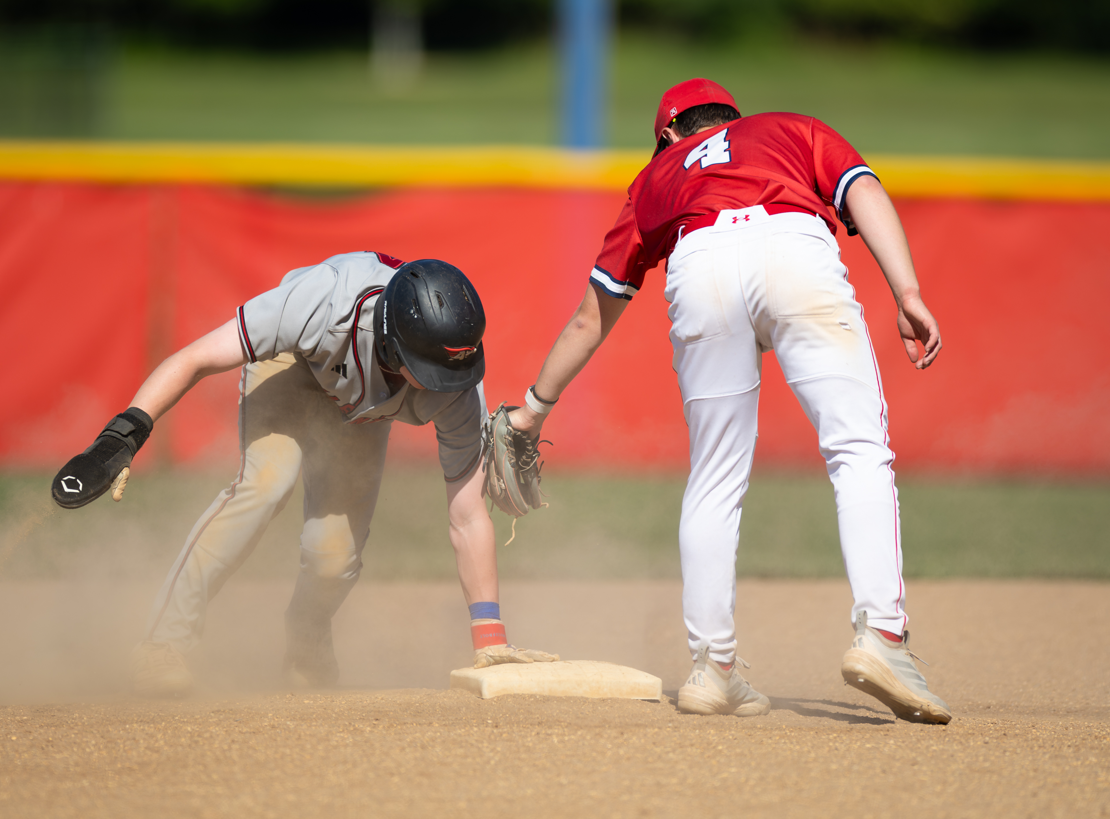 A baseball player slides into a base while another player in red reaches for him, with a red outfield fence and green grass in the background.