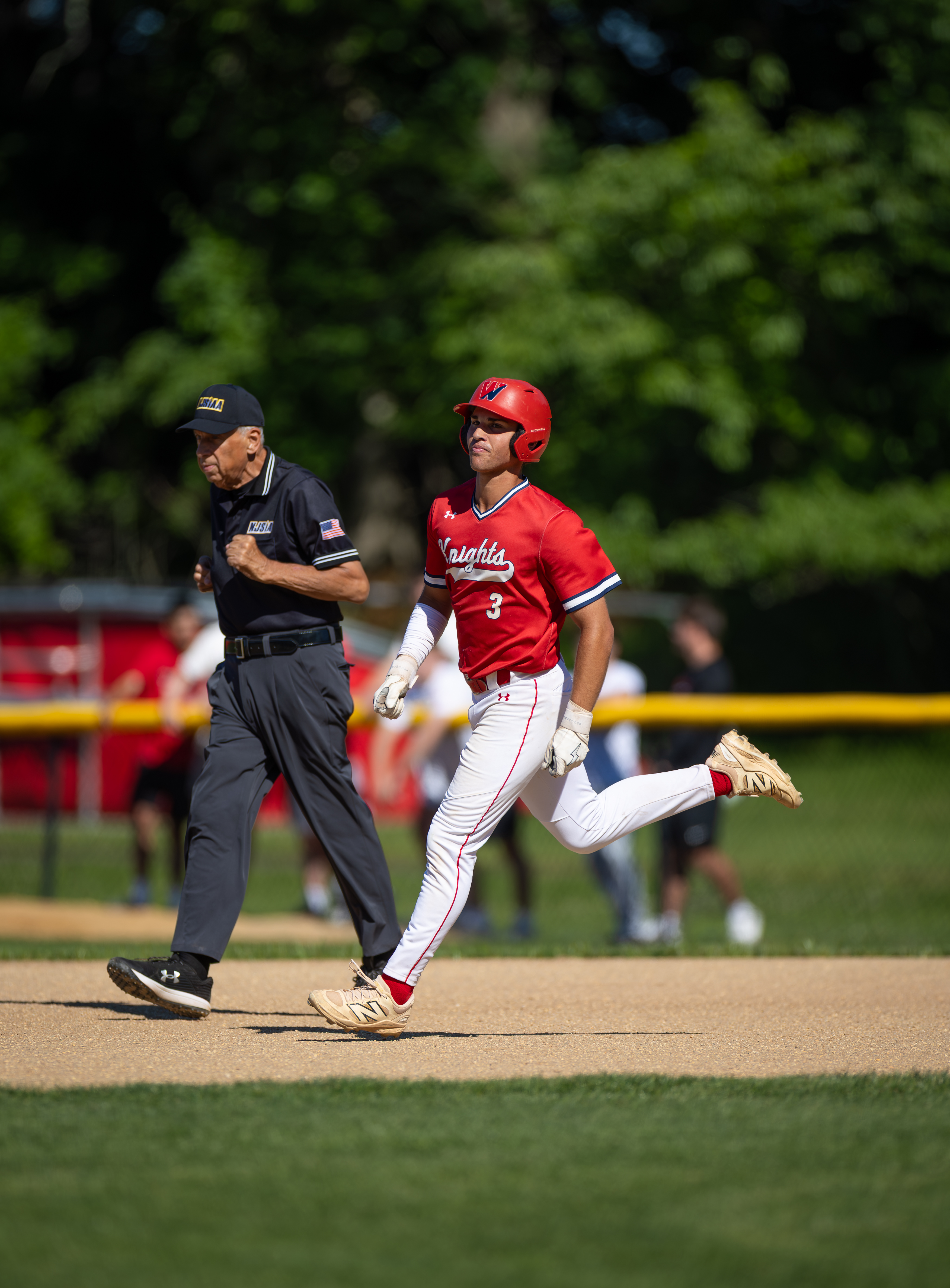 A baseball player in a red uniform runs on a dirt path in the foreground, while an umpire walks behind him, with a blurred background of trees and a fence.