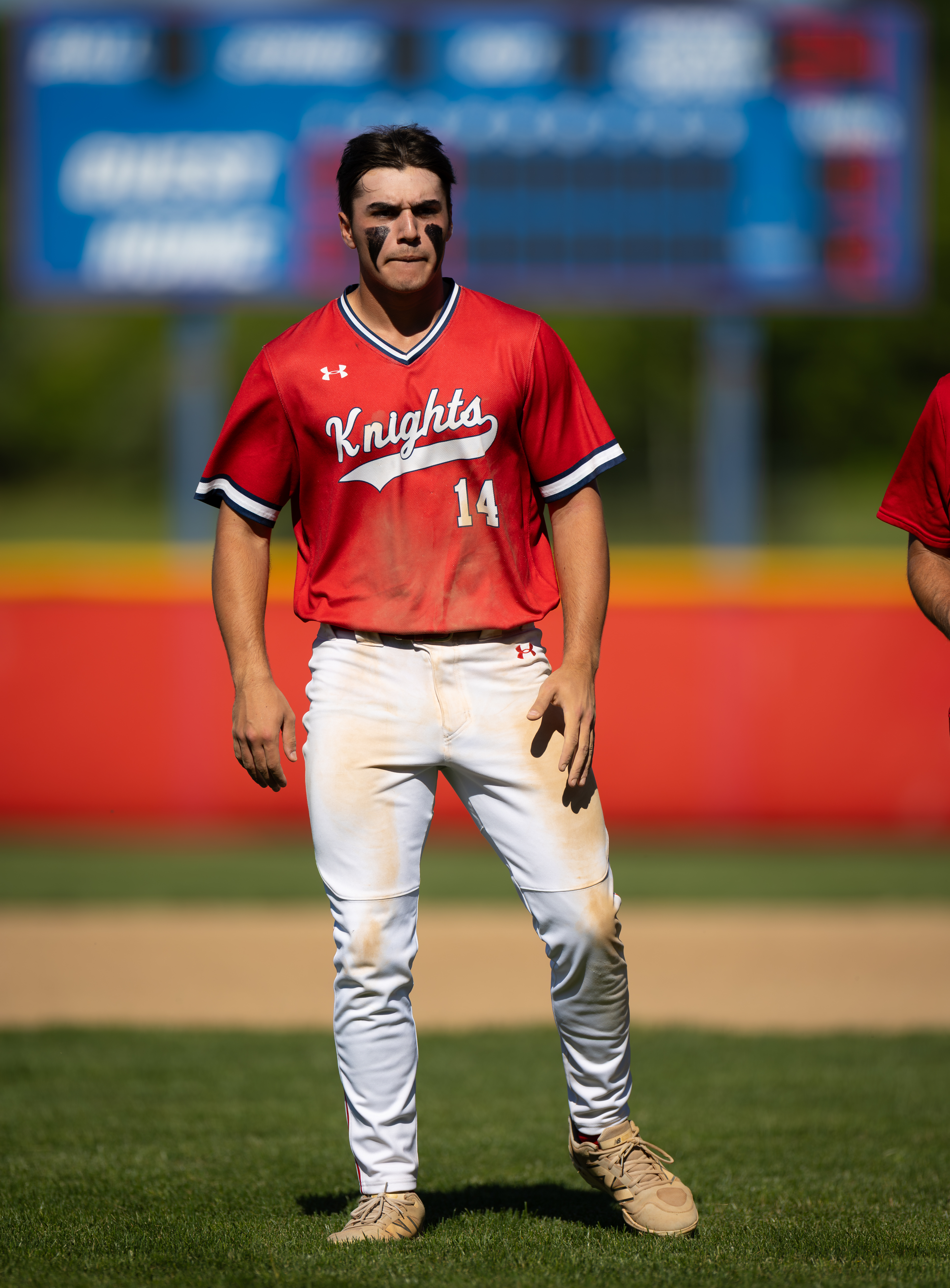 A baseball player in a red "Knights" jersey stands on a grassy field with a blurred scoreboard in the background.
