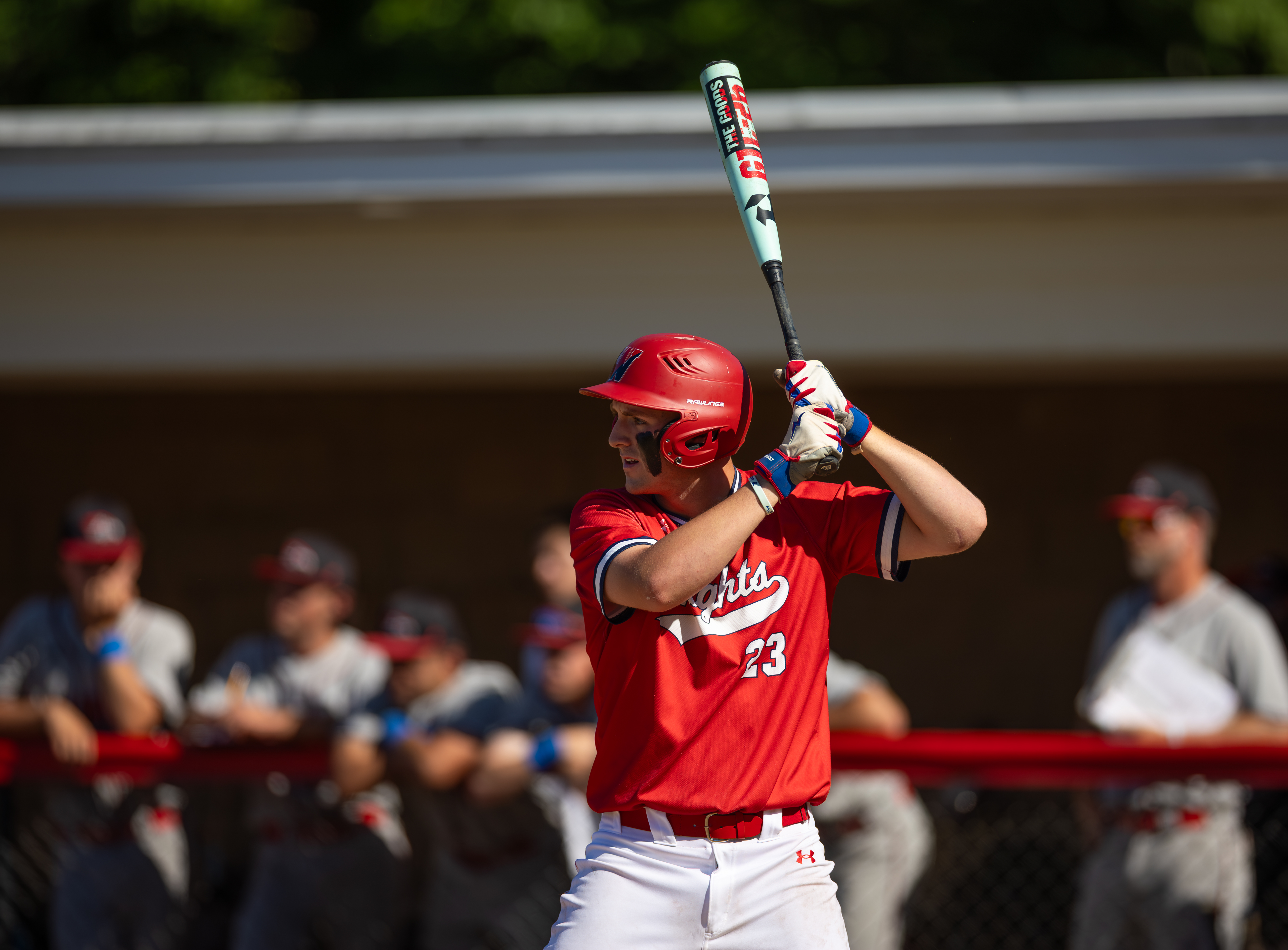 A baseball player in a red uniform with the number 23 on his jersey stands at bat, holding a light blue and white bat, with a dugout full of teammates and a blurred background of trees and a building.