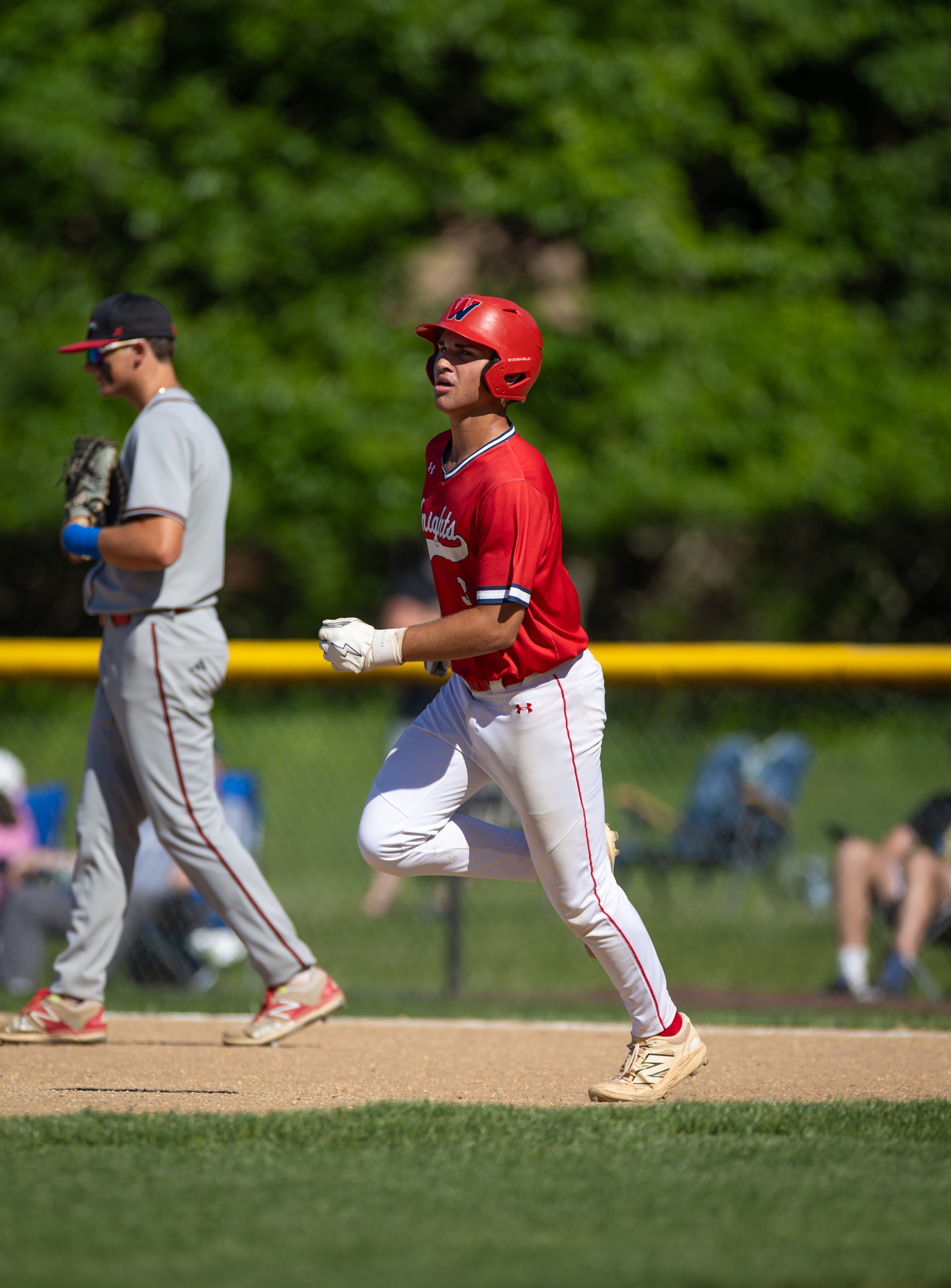 A young baseball player in a red uniform runs on a dirt field in the foreground, with another player and blurred spectators in the background under a tree-lined sky.