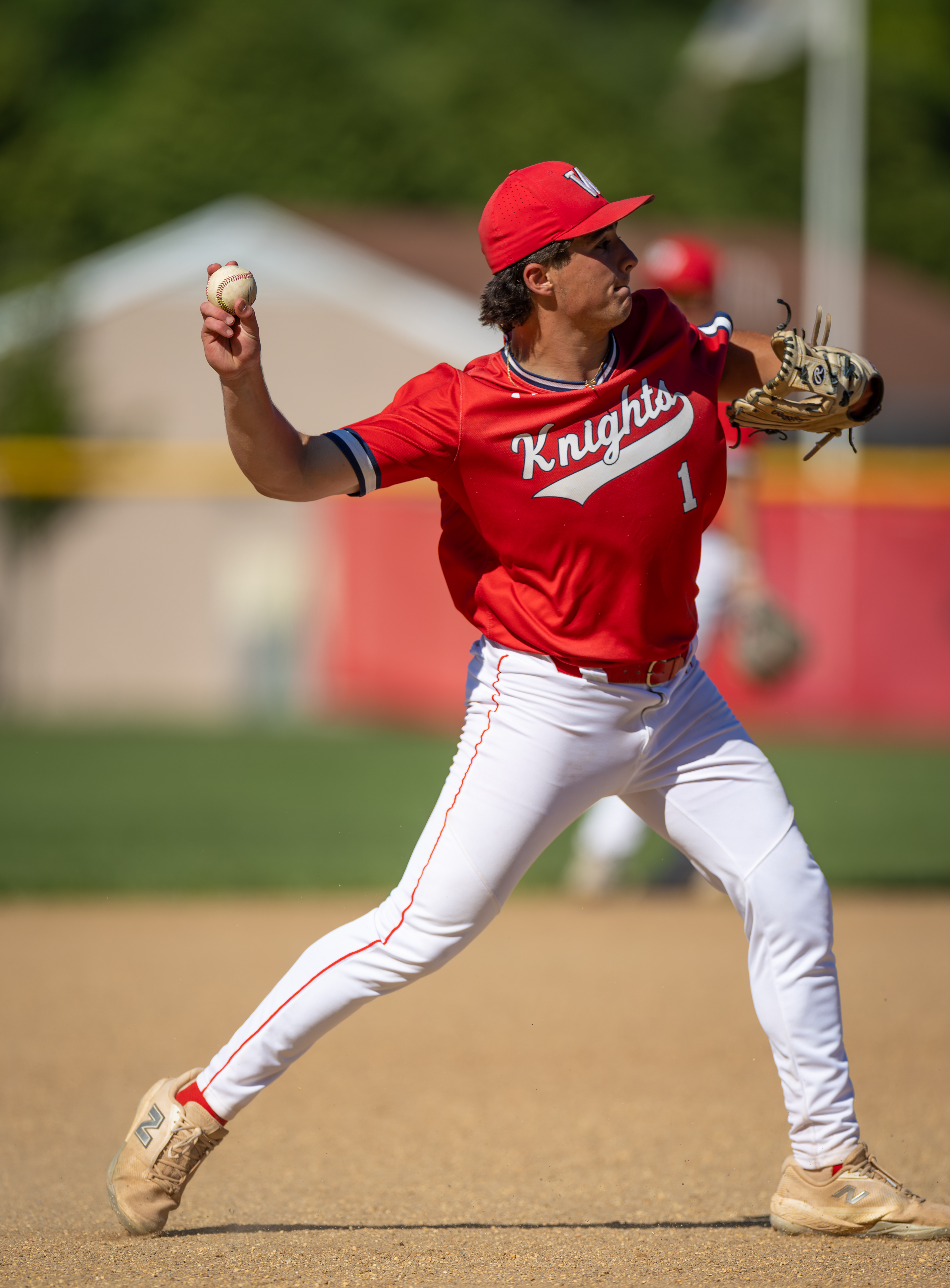 A baseball player in a red uniform prepares to throw a ball in the foreground, with a blurred background of a baseball field and buildings.
