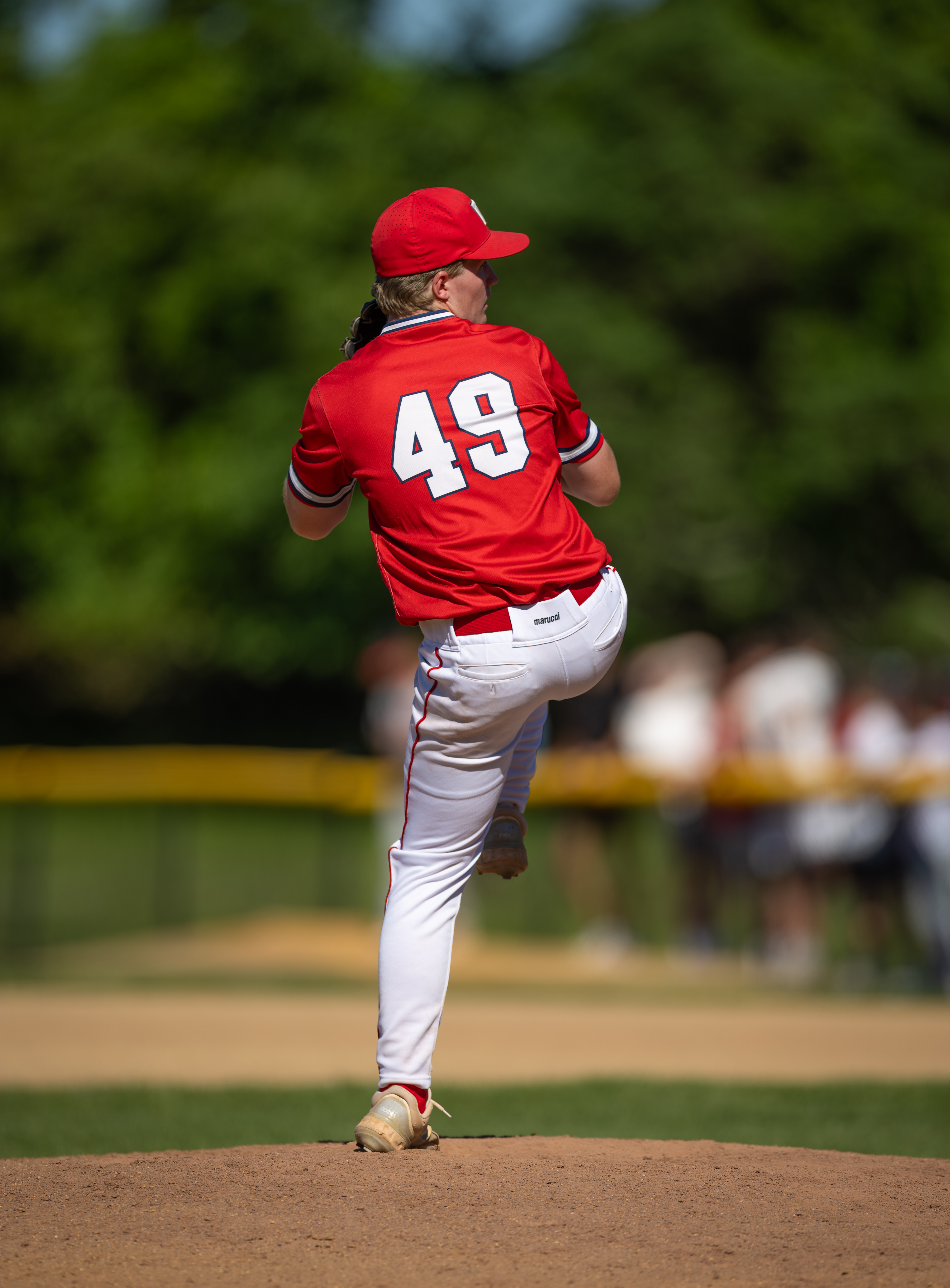 A baseball pitcher in a red jersey with the number 49 on his back stands on the mound, with blurred figures and green trees in the background.