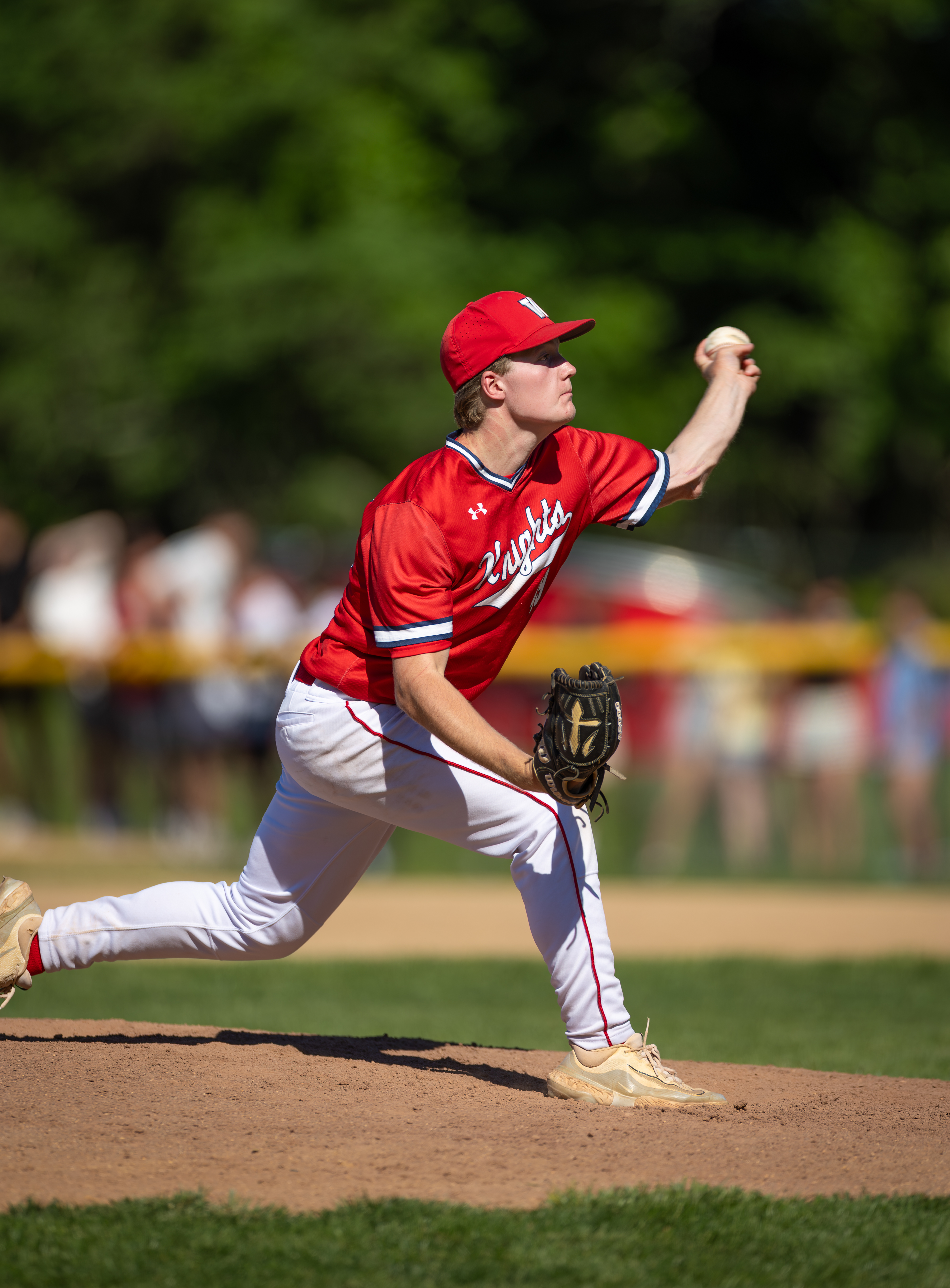 A baseball pitcher in a red uniform throws a ball on a dirt mound, with blurred spectators and green trees in the background.