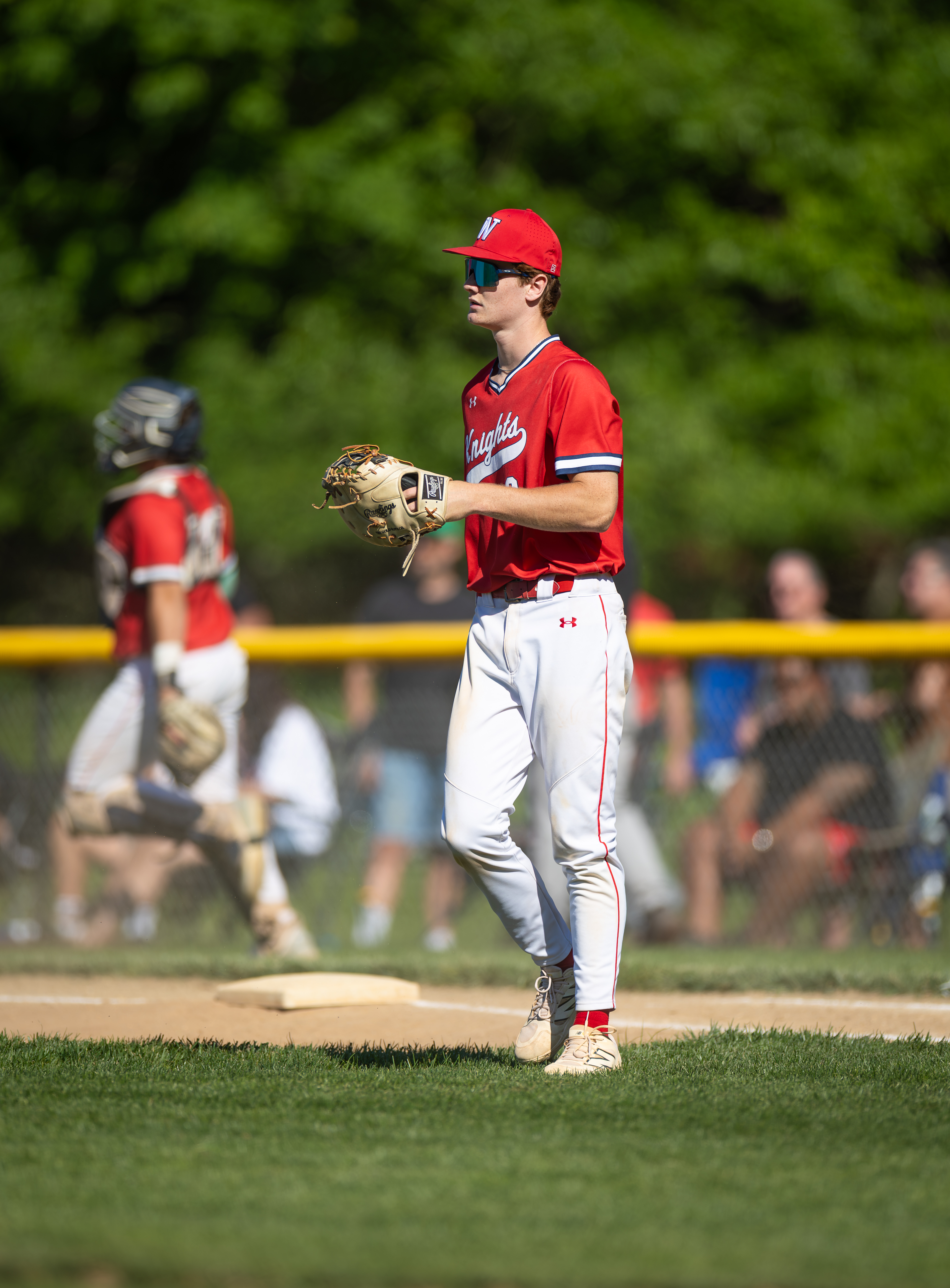 A baseball player in a red uniform stands on a dirt infield with a catcher in the background, while blurred spectators watch from behind a yellow fence under a canopy of green trees.