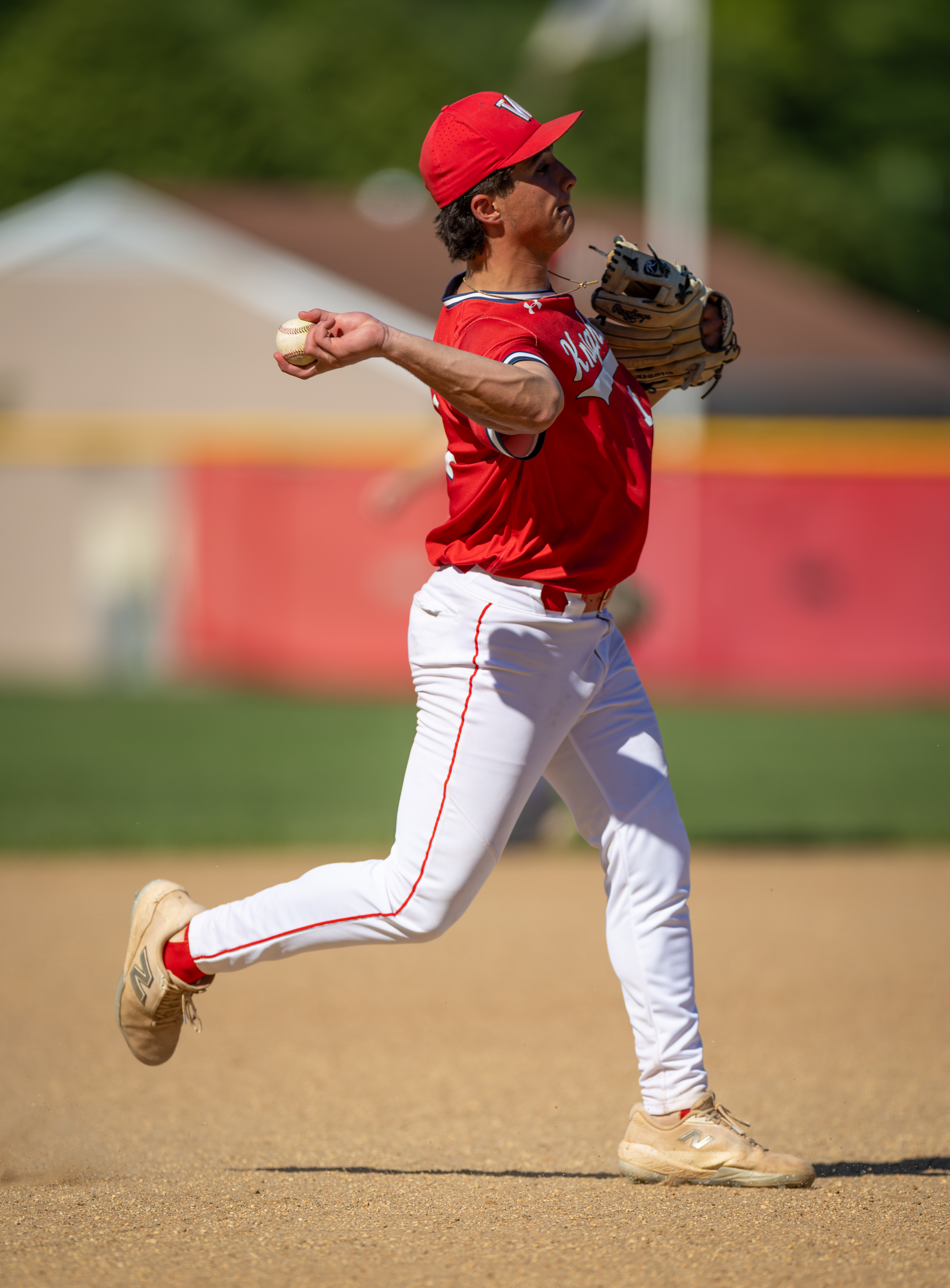 A baseball player in a red uniform throws a ball on a dirt field with a blurred background of a fence and trees.
