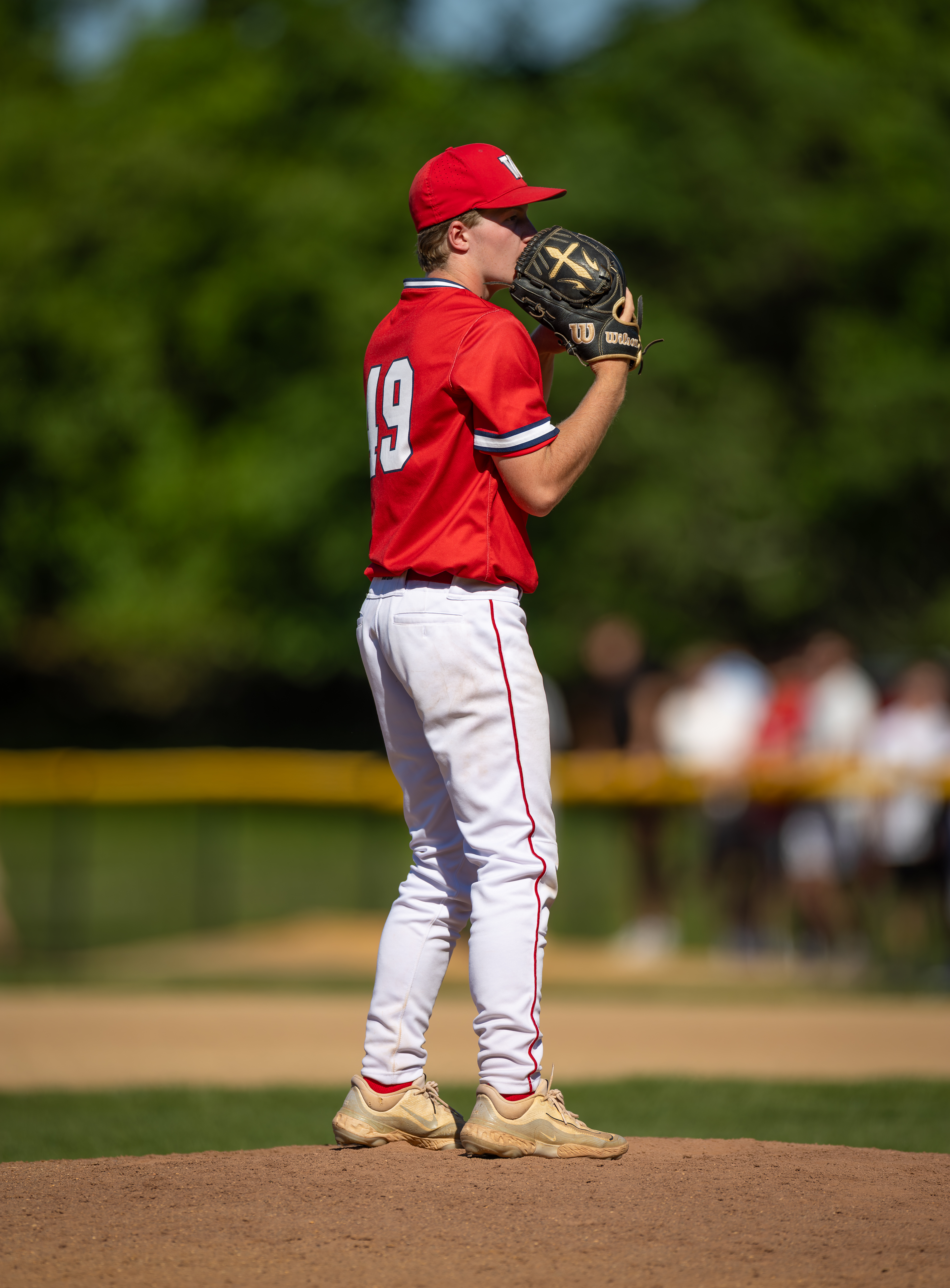 A baseball pitcher in a red jersey and white pants stands on the mound, with blurred spectators and green trees in the background.