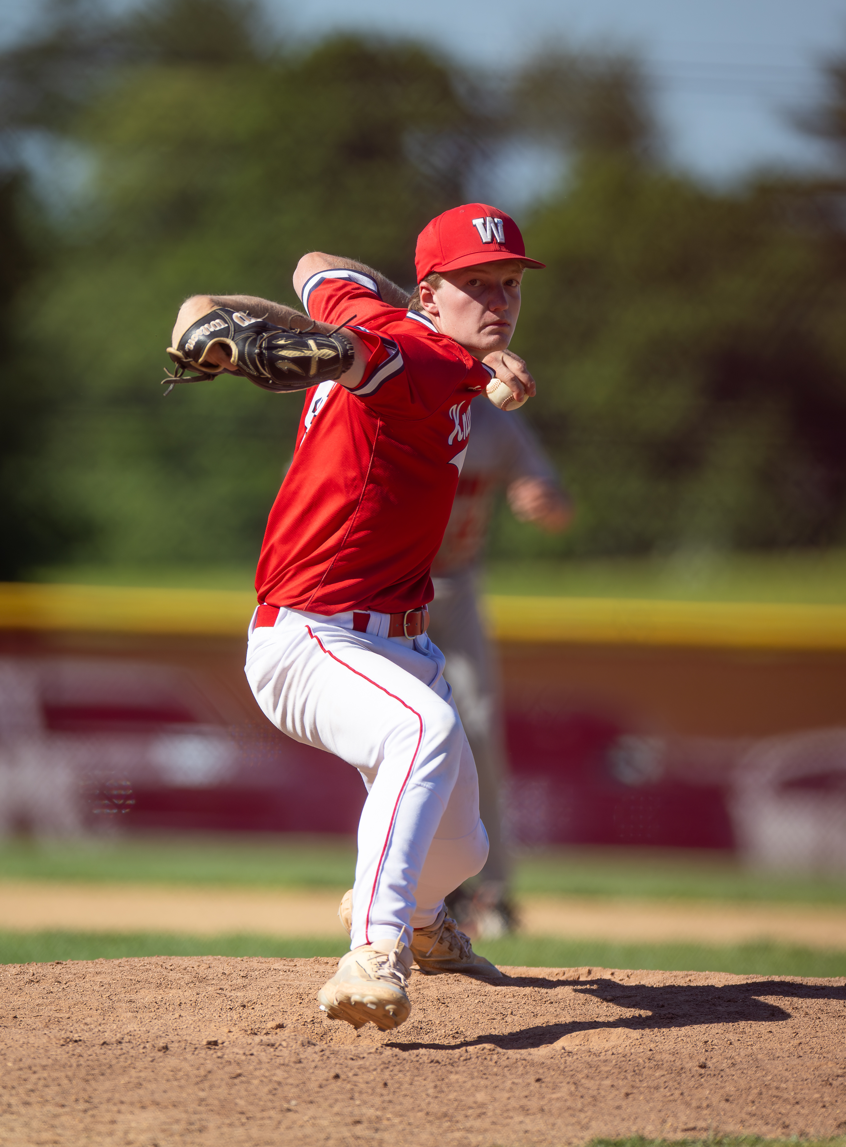 A baseball pitcher in a red uniform throws a pitch on a dirt mound, with a blurred outfield and yellow fence in the background.
