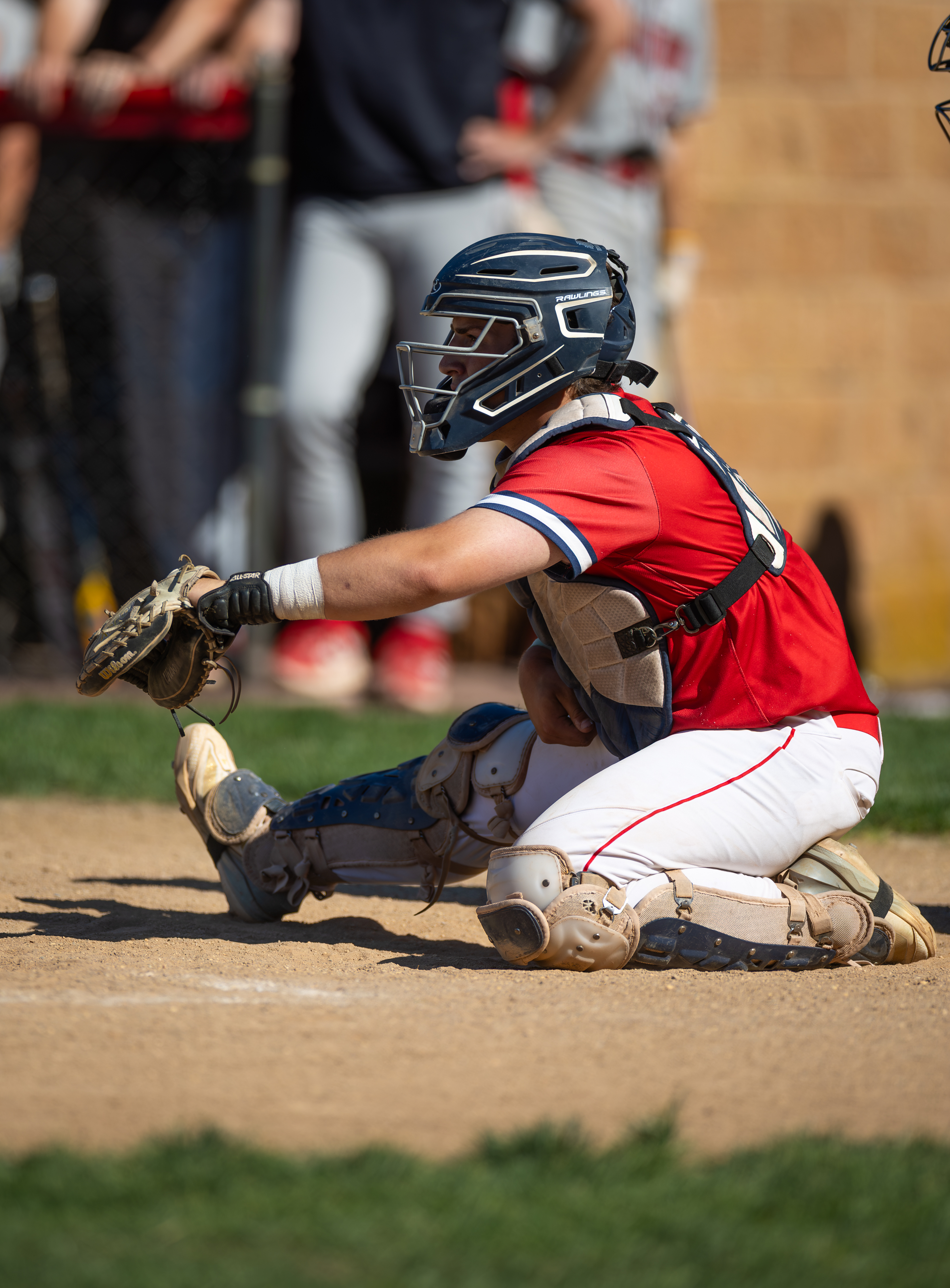 A baseball catcher in red and white uniform is crouched on the dirt infield, with blurred figures in the background.
