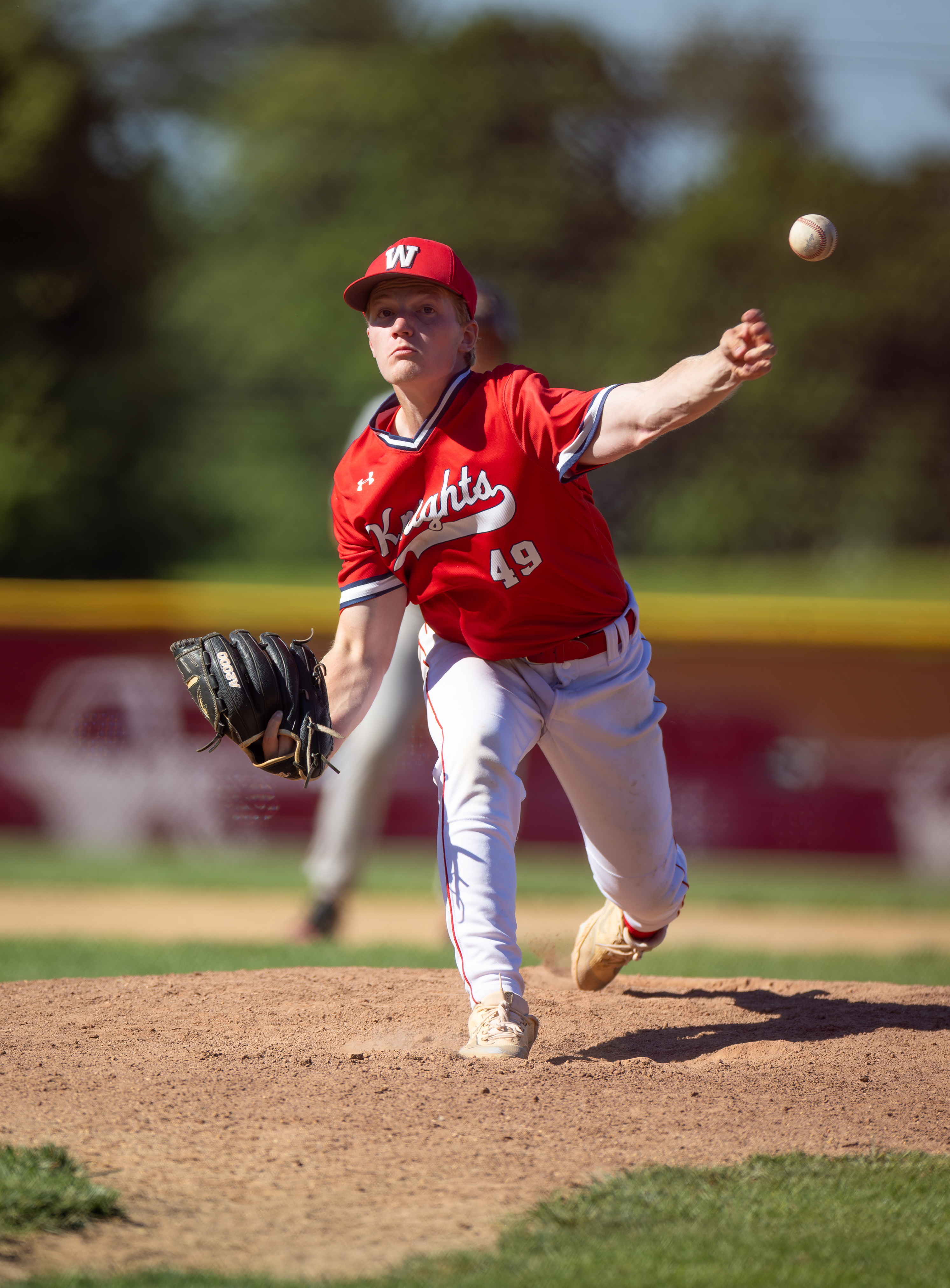 A young baseball pitcher in a red uniform with "Knights" and the number 49 on it throws a ball on a dirt mound, with a blurred baseball field and trees in the background.