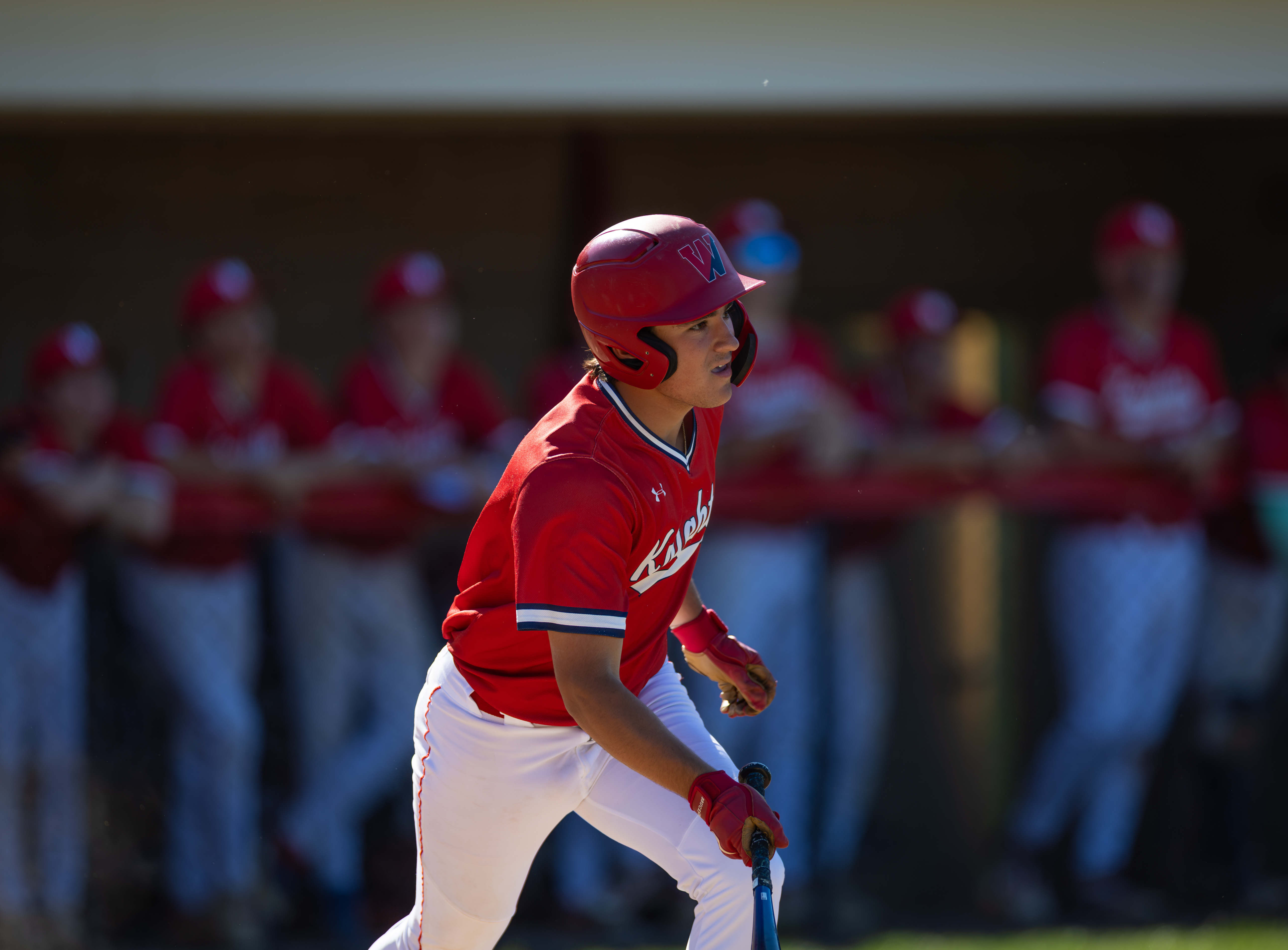 A young baseball player in a red uniform runs in the foreground with his teammates blurred in the background.