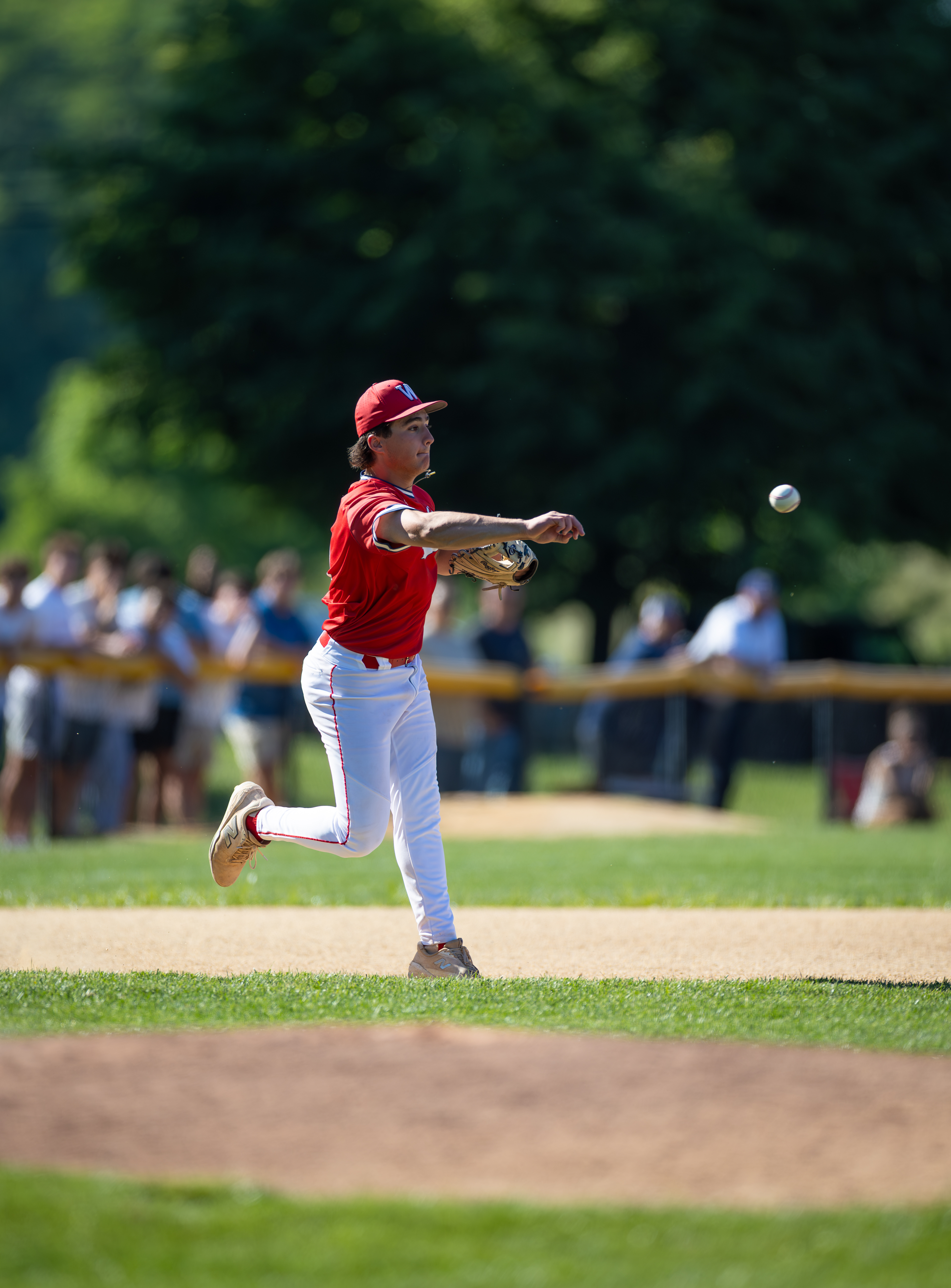 A young baseball player in a red uniform throws a ball on a sunny field with spectators blurred in the background.