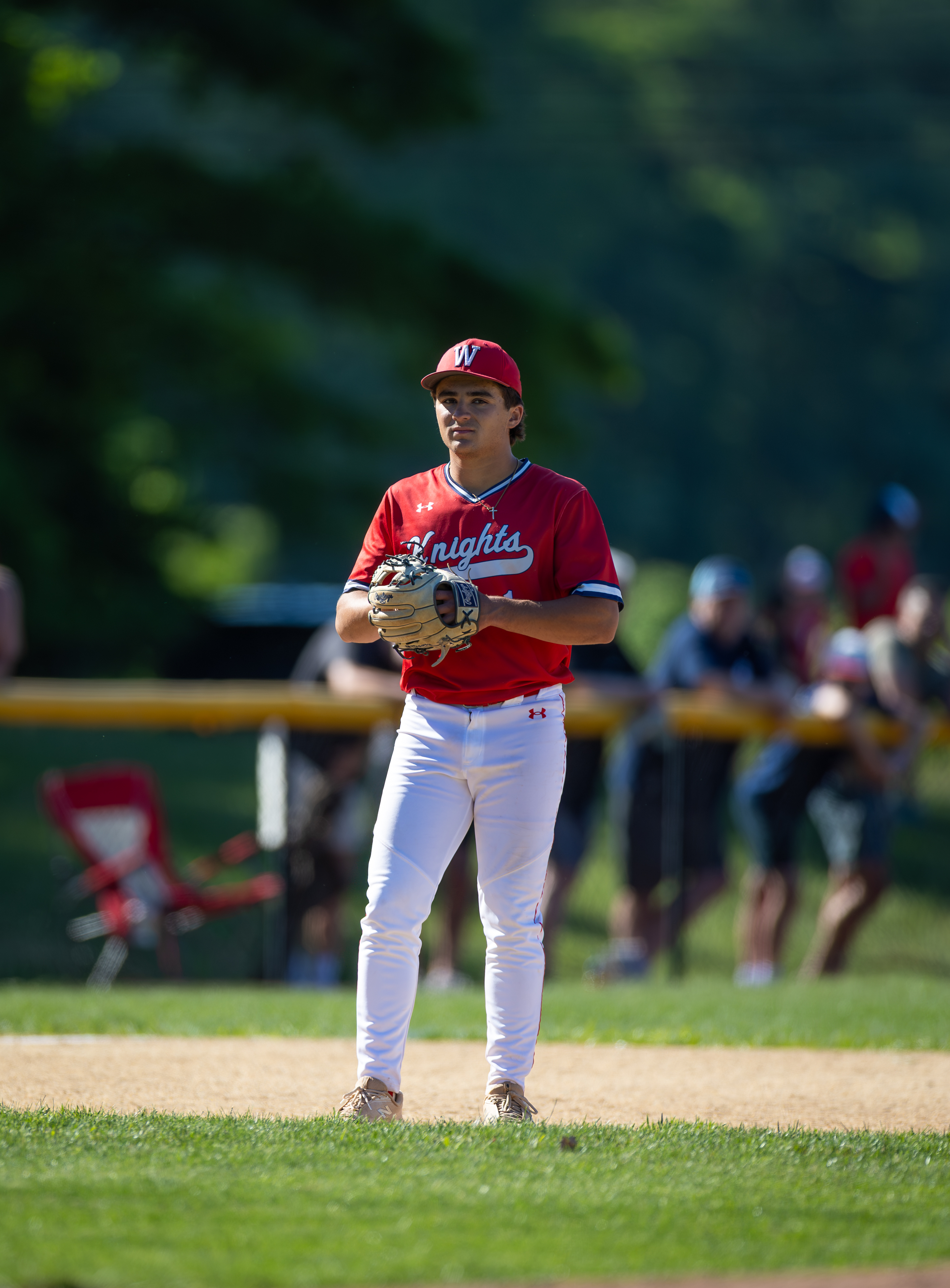 A young baseball player in a red uniform stands on a grassy field with a blurred background of spectators and trees.