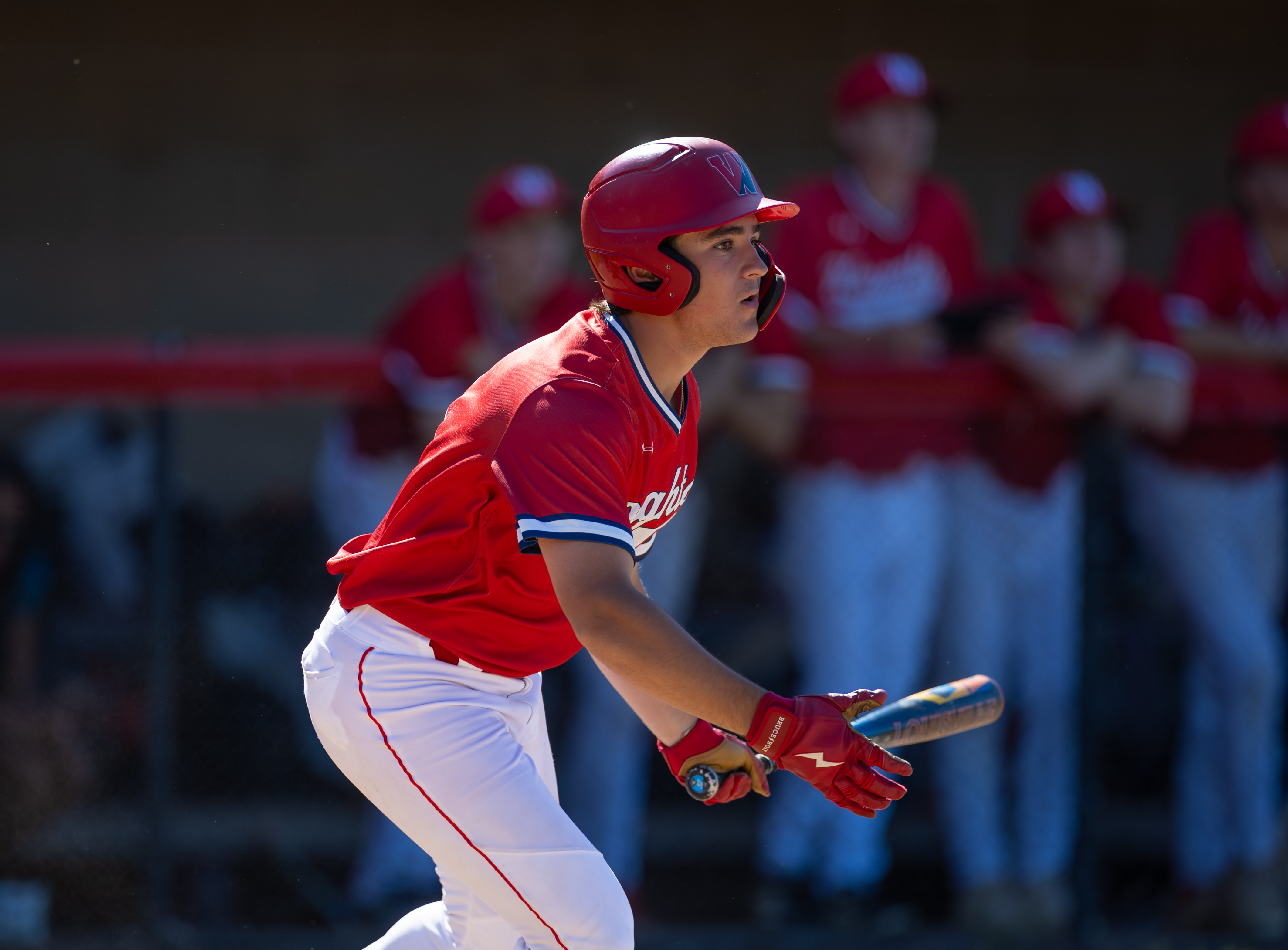 A baseball player in a red uniform swings a bat in the foreground, with blurred figures in red uniforms in the background.