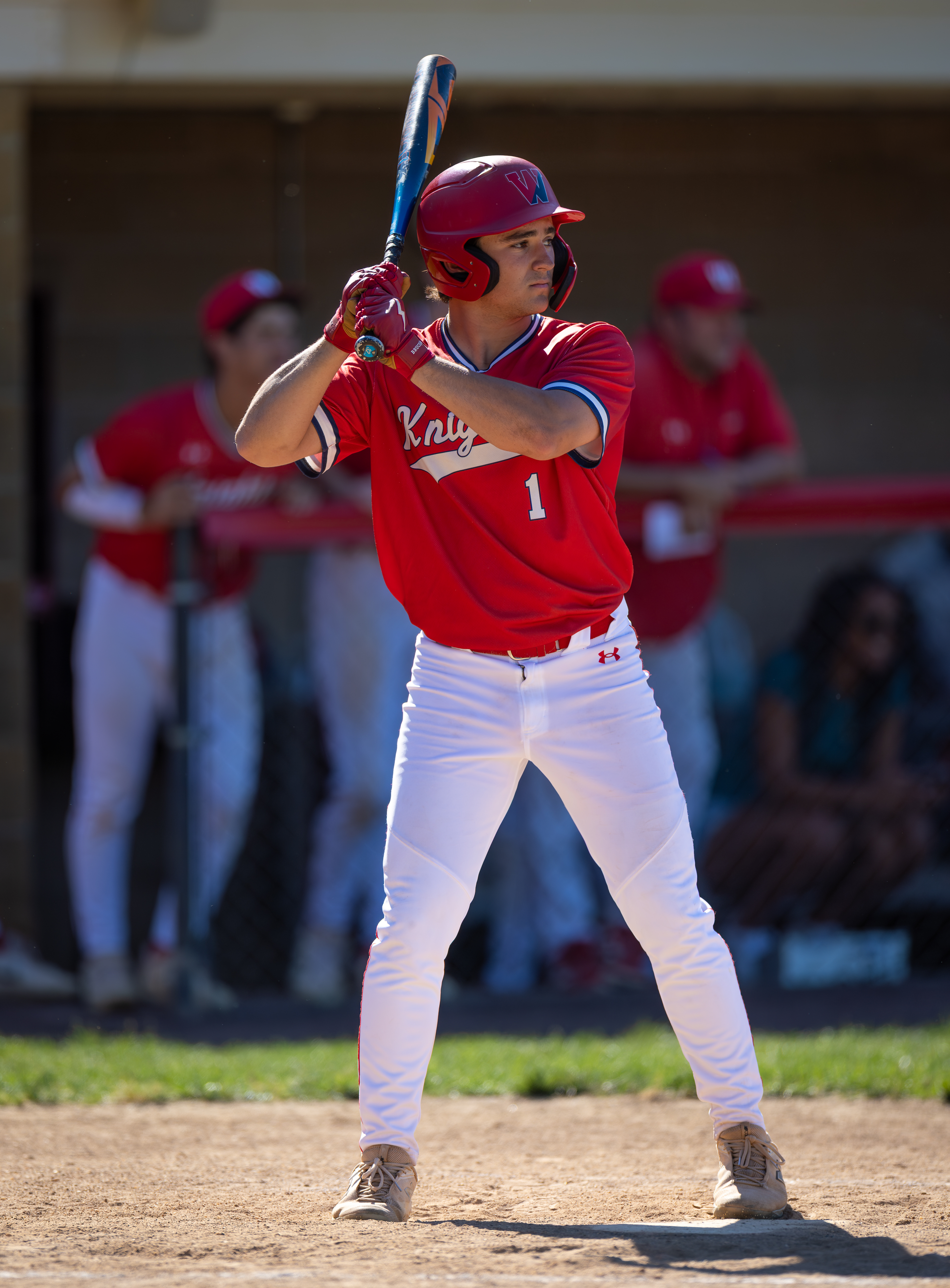 A baseball player in a red uniform with the number 1 on his back stands at home plate, ready to bat, with teammates and spectators blurred in the background.