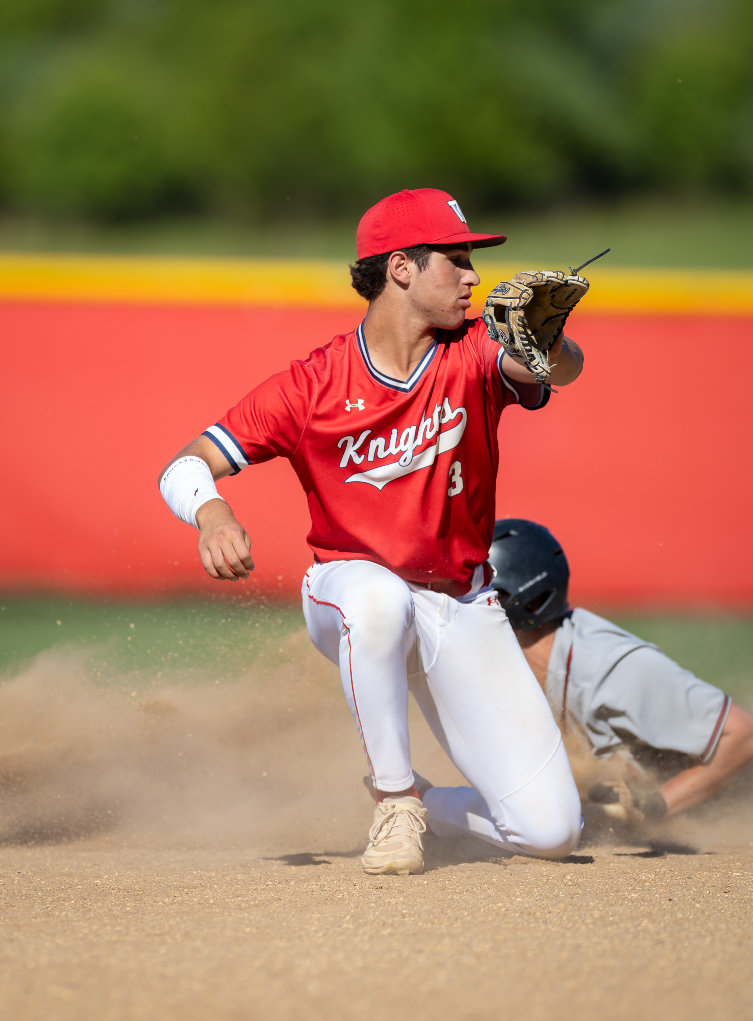 A baseball player in a red uniform slides into a base as another player attempts to tag him out, with a blurred red outfield wall in the background.