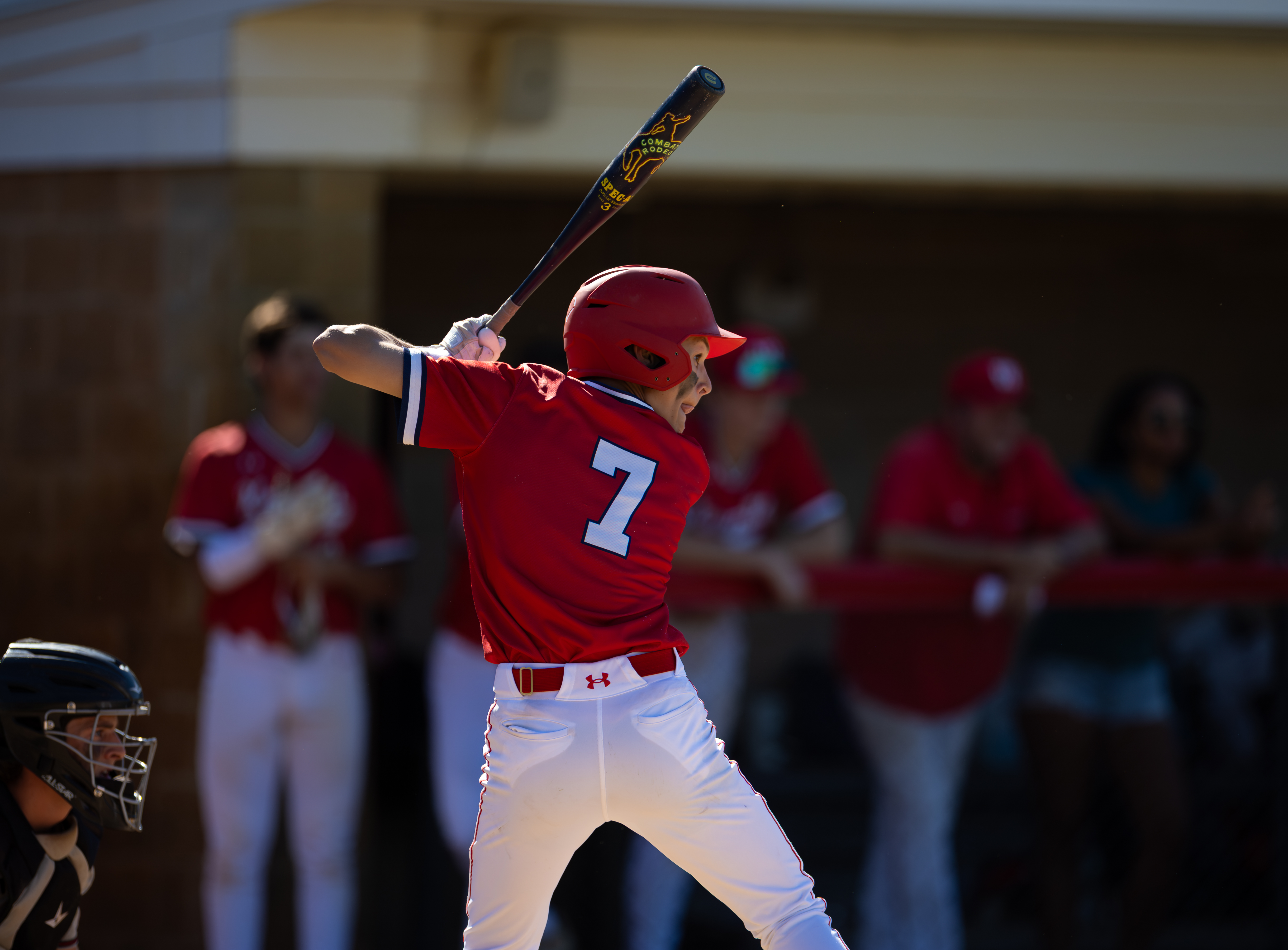 A young baseball player in a red uniform with the number 7 on his back swings a bat in the foreground, while blurred figures in red uniforms and a dugout are visible in the background.