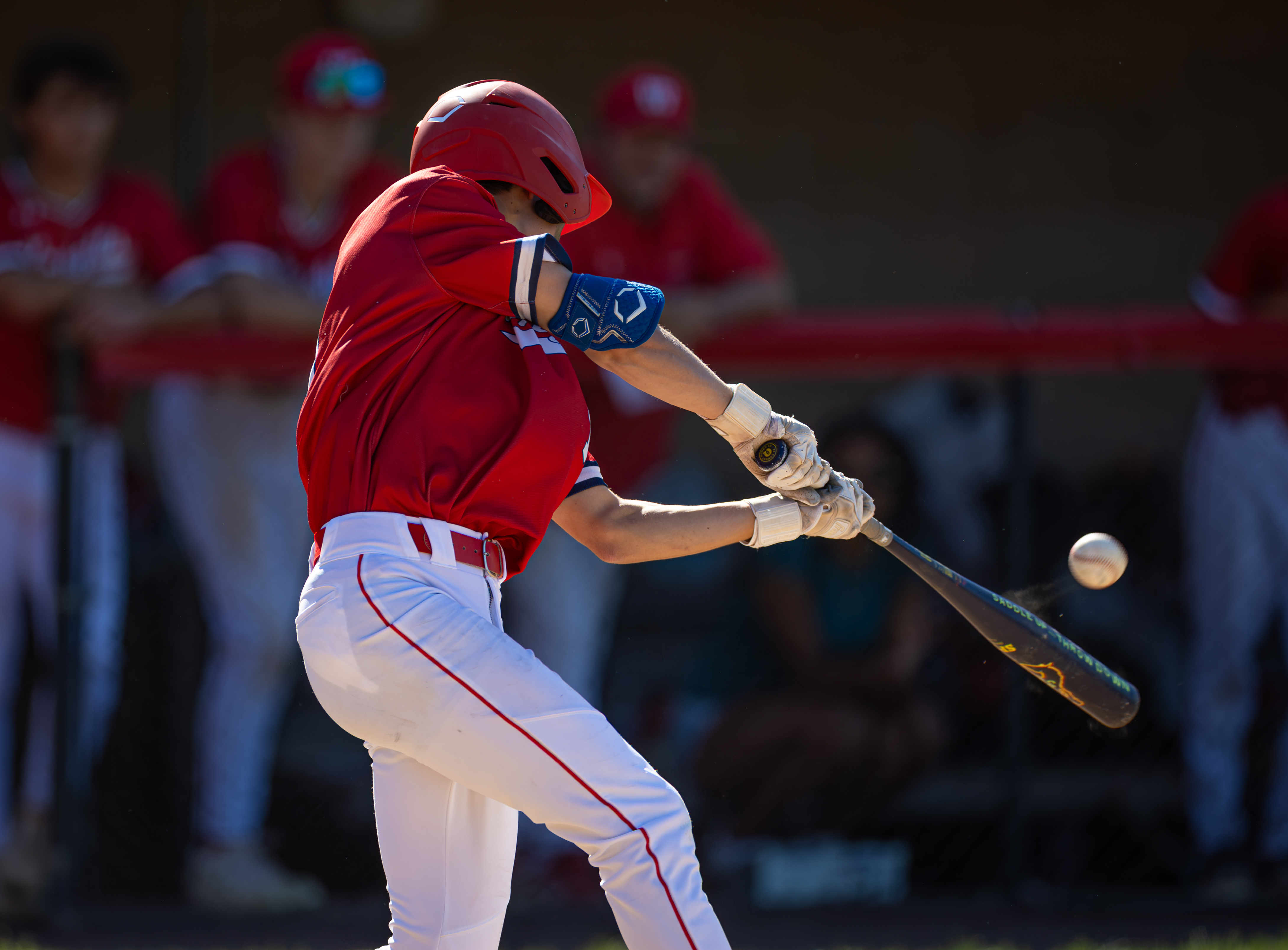 A baseball player in a red uniform swings a bat at a ball in the foreground, with blurred figures in red uniforms in the background.