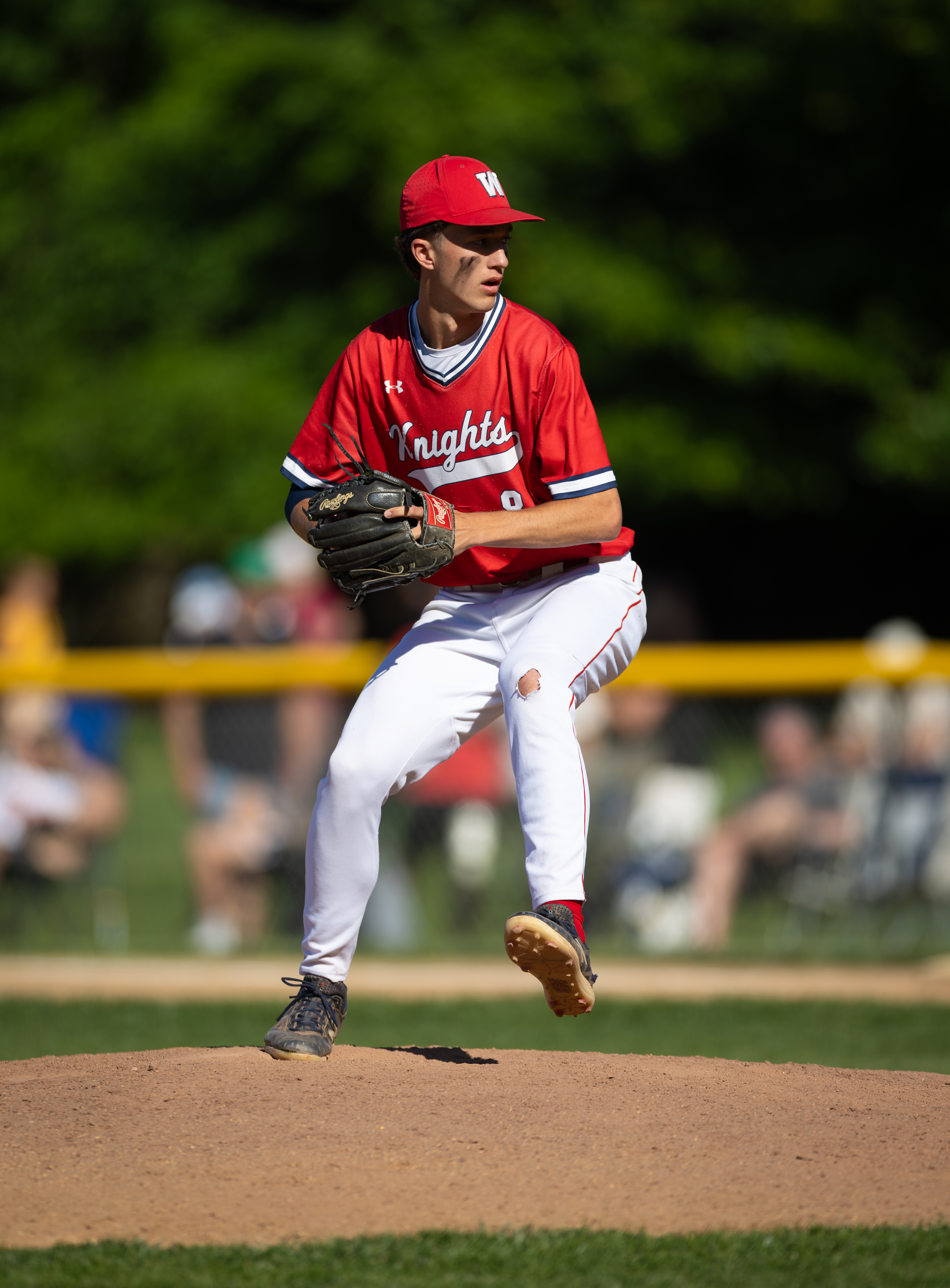 A young baseball pitcher in a red uniform is in mid-throw on a dirt mound, with blurred spectators and green trees in the background.