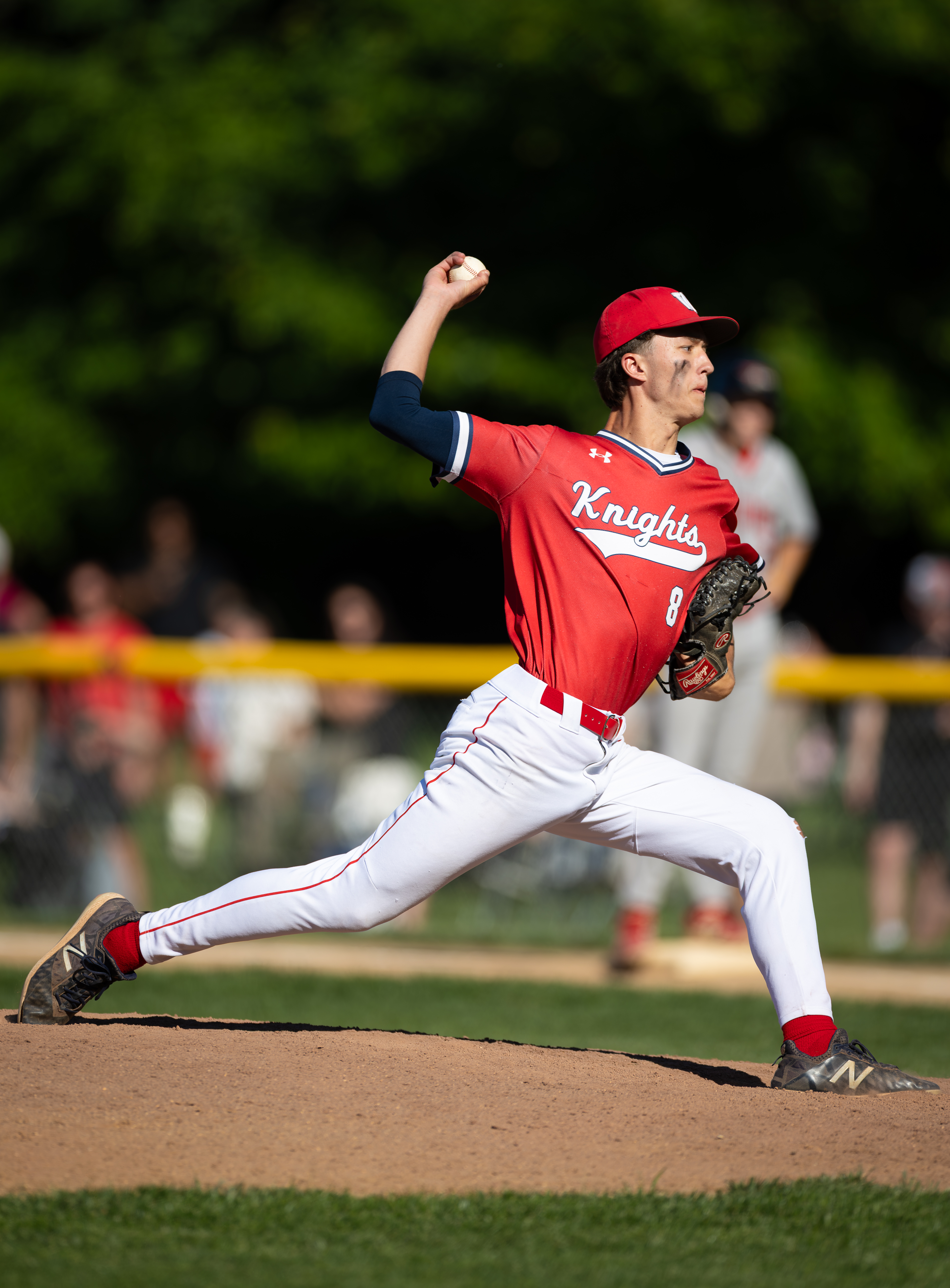 A baseball pitcher in a red Knights uniform throws a pitch on a dirt mound, with blurred spectators and trees in the background.
