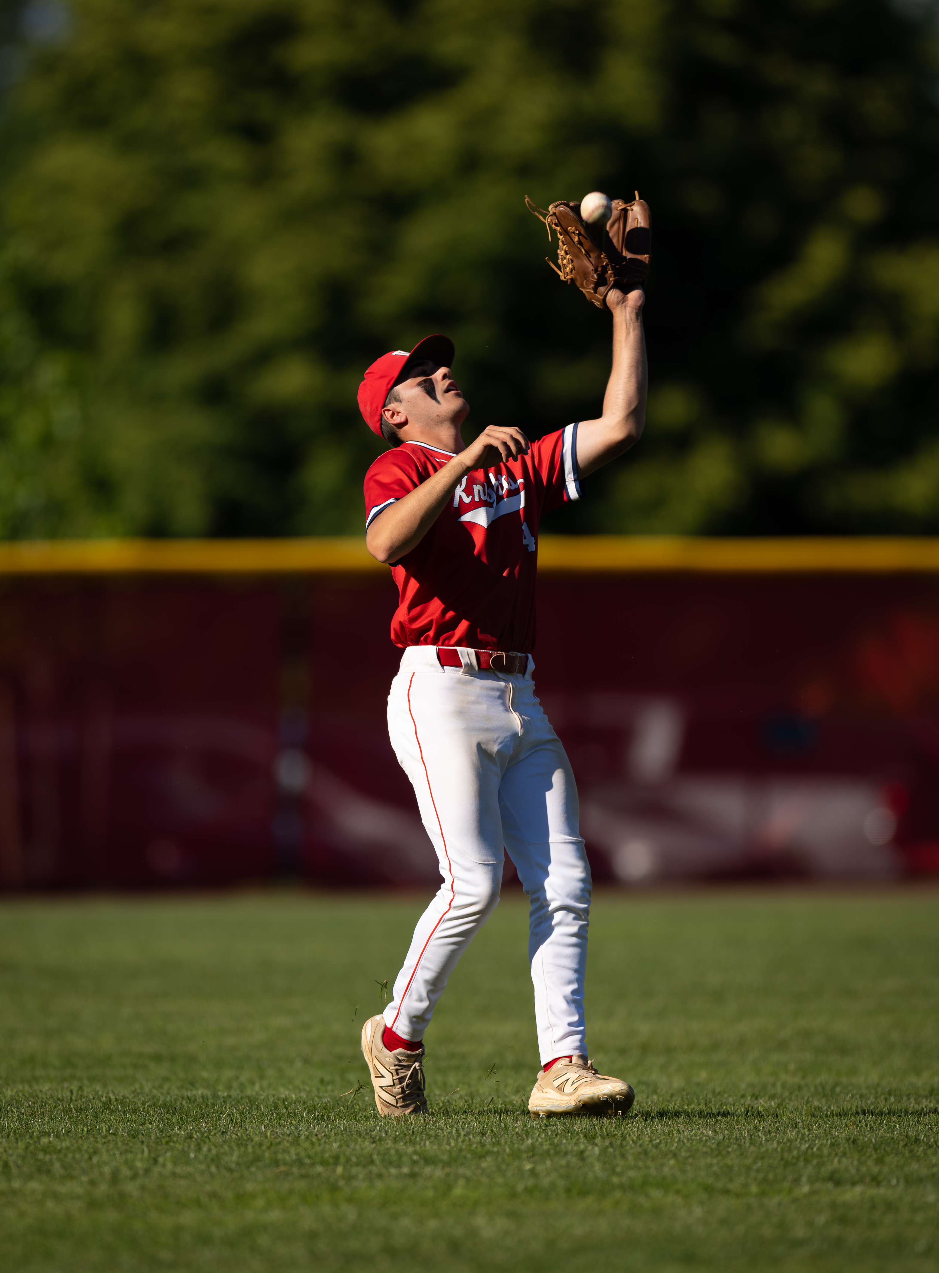 A baseball player in a red uniform catches a fly ball in a grassy field with a blurred green tree line in the background.