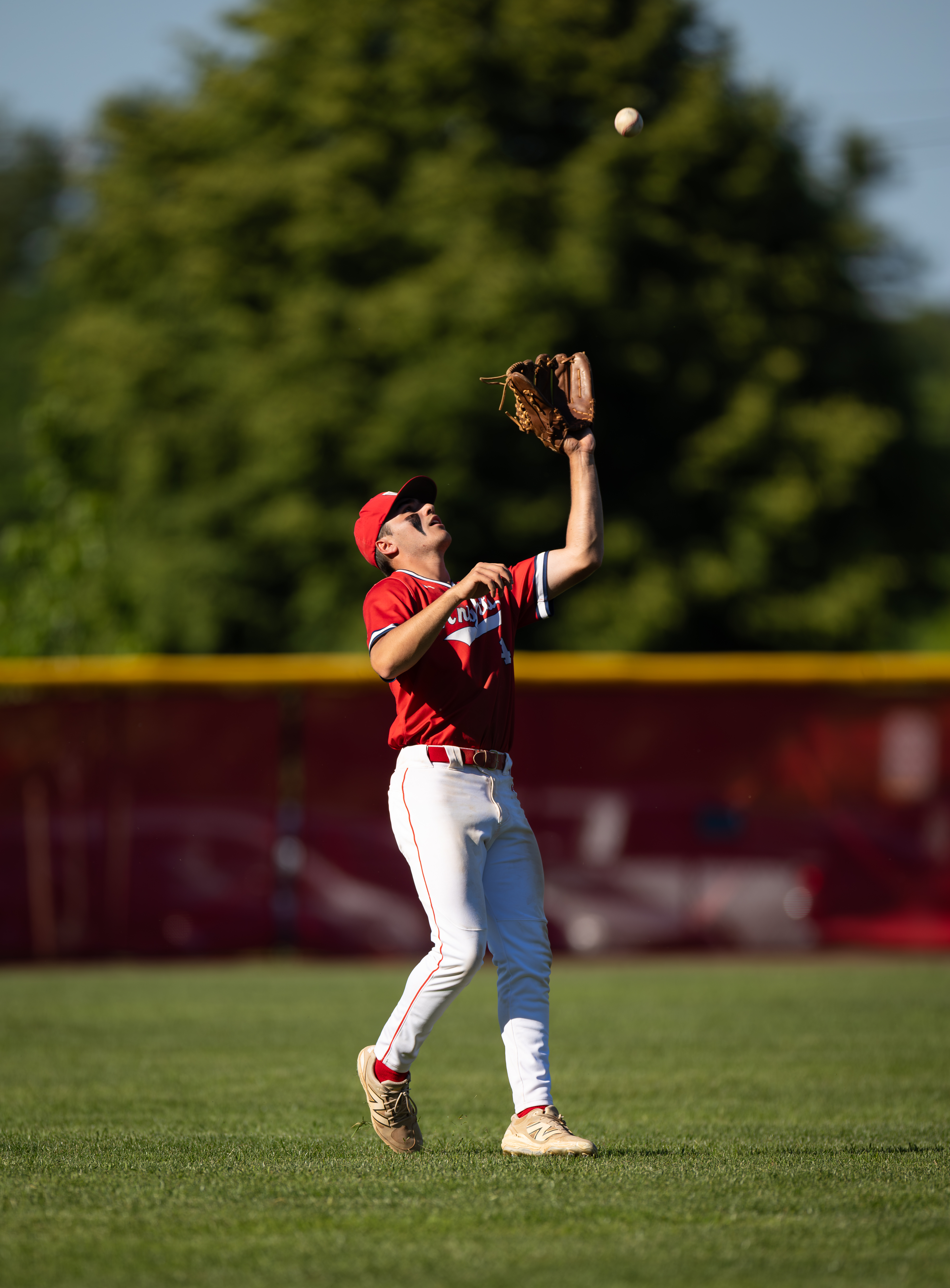 A baseball player in a red uniform reaches up to catch a ball in the foreground, with a blurred green tree line and a red fence in the background.