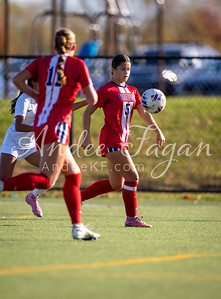 Two soccer players in red uniforms are on a green field, with a blurred background of trees and a car.