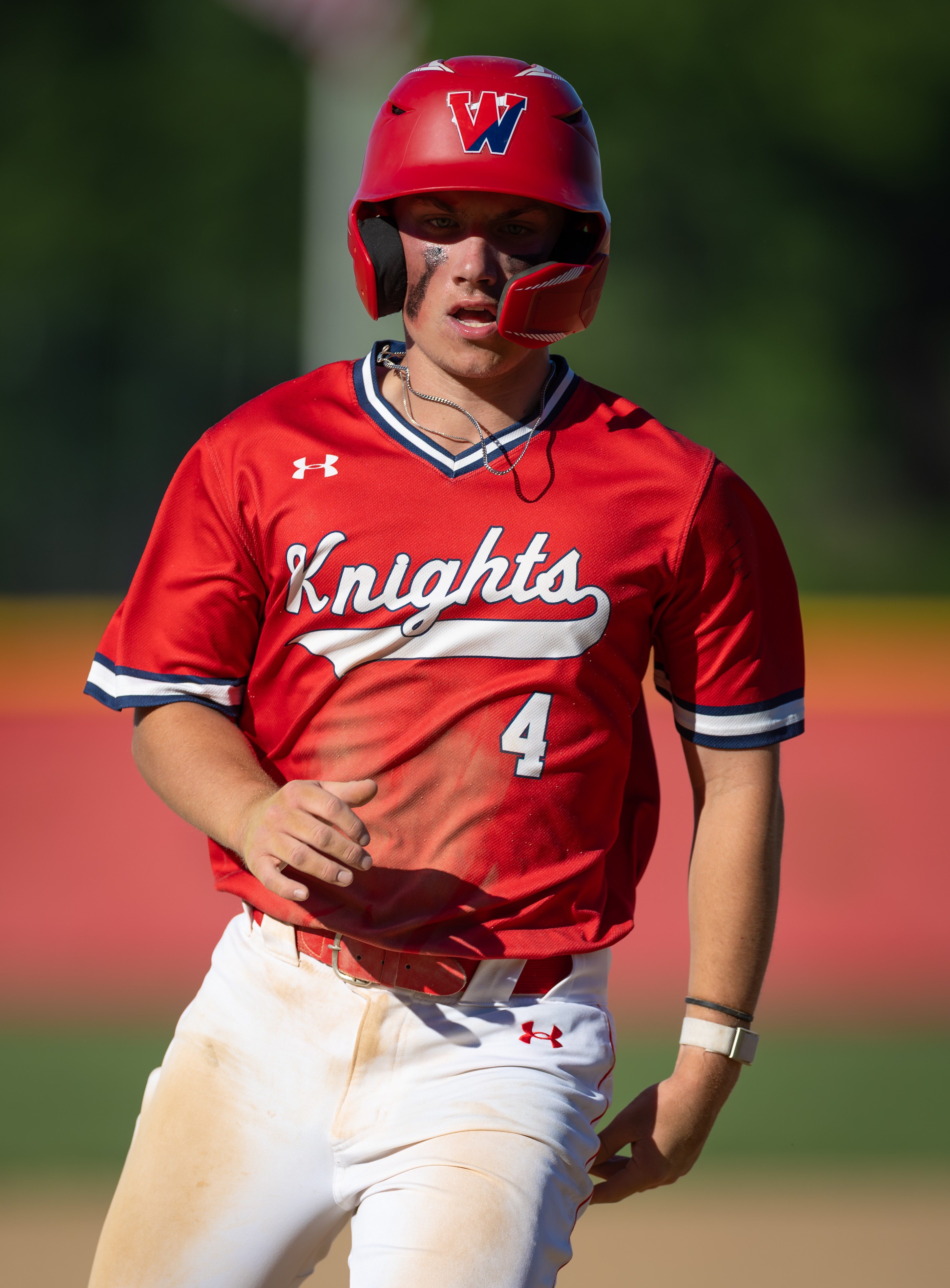 A young baseball player in a red "Knights" jersey and helmet stands on a dirt field with a blurred green and red background.