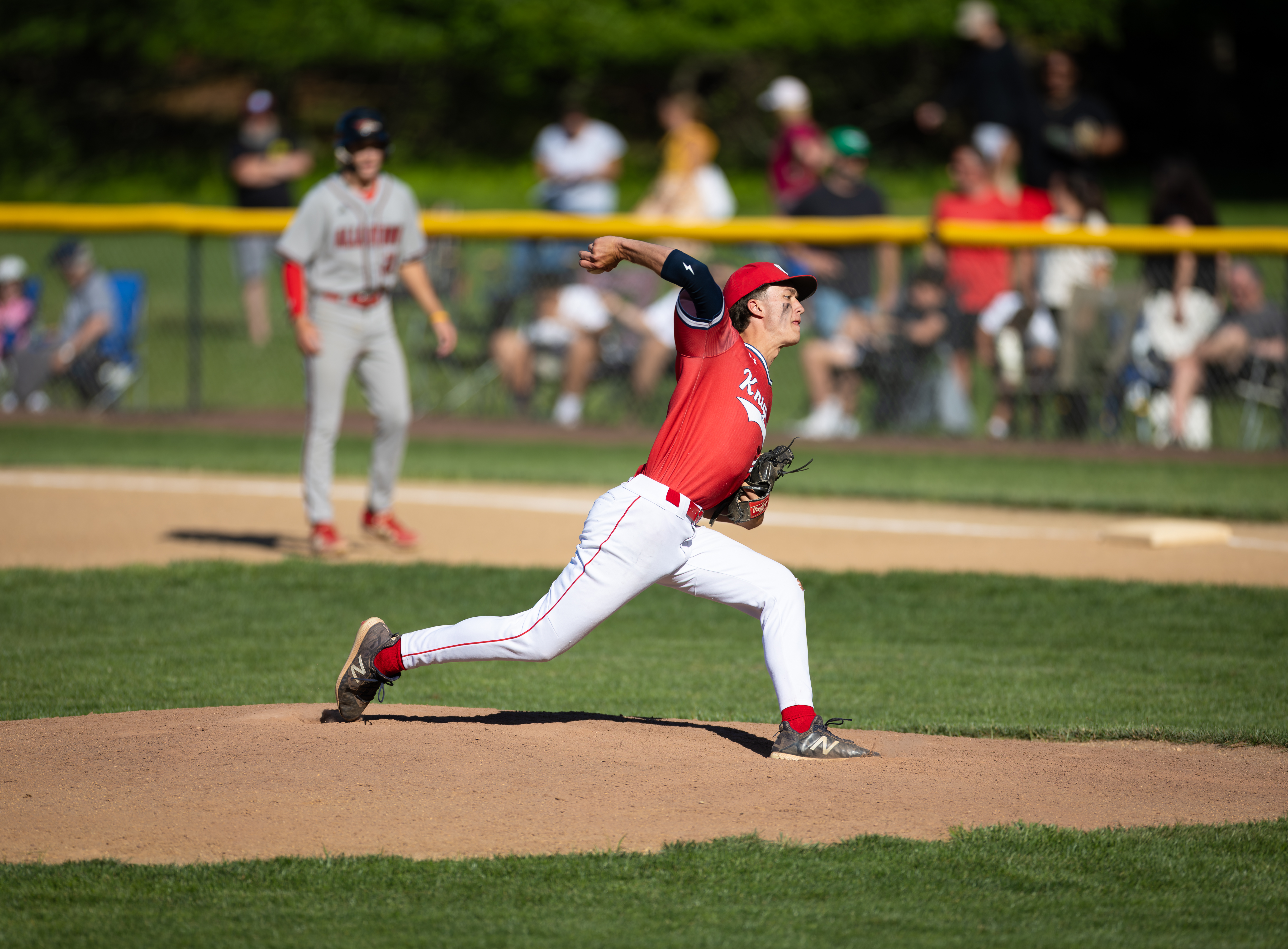 A baseball pitcher in a red uniform throws a pitch on a dirt mound in the foreground, with blurred spectators and trees in the background.