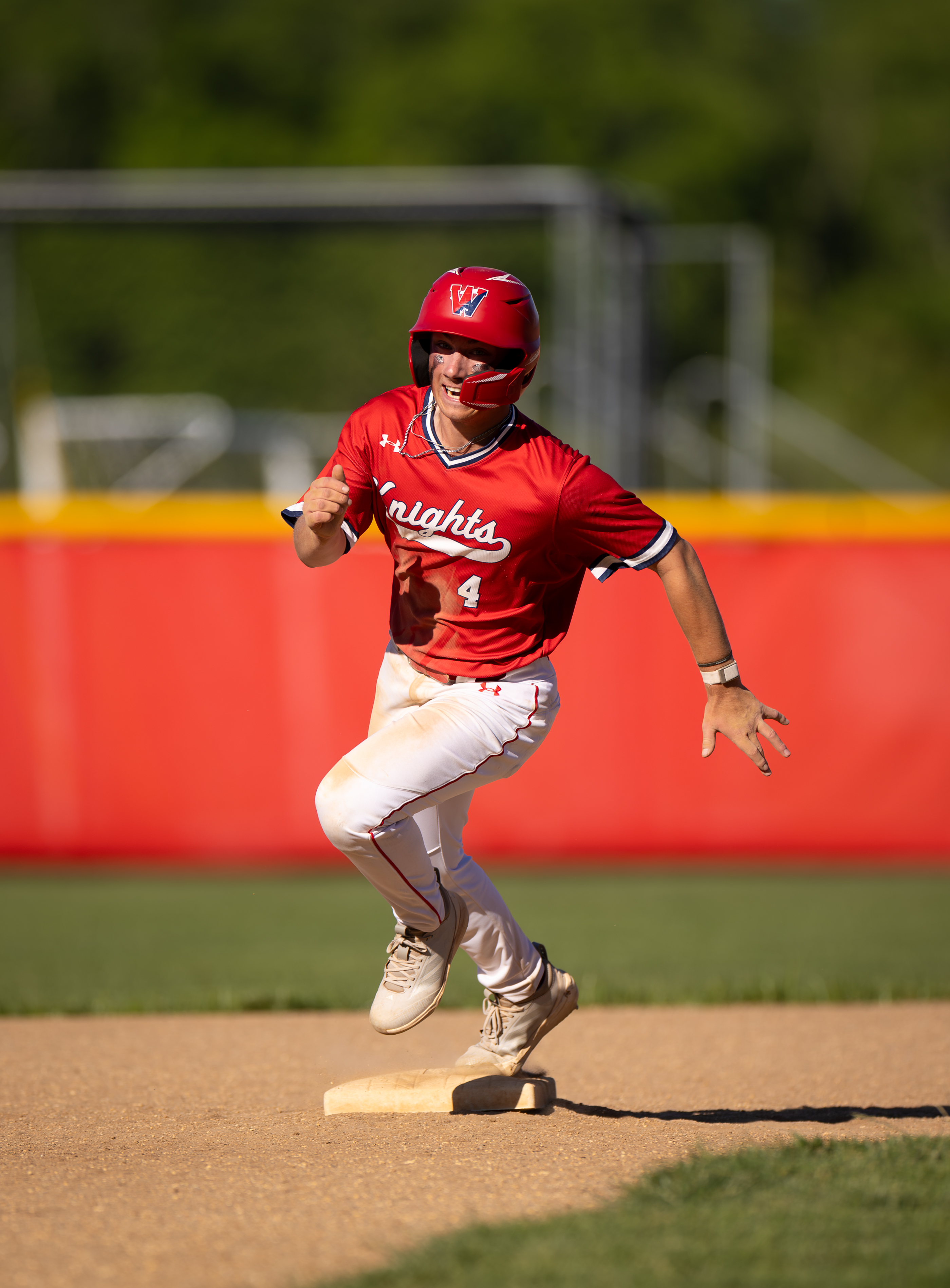 A baseball player in a red uniform rounds a base in the foreground, with a bright red outfield fence and green trees in the background.