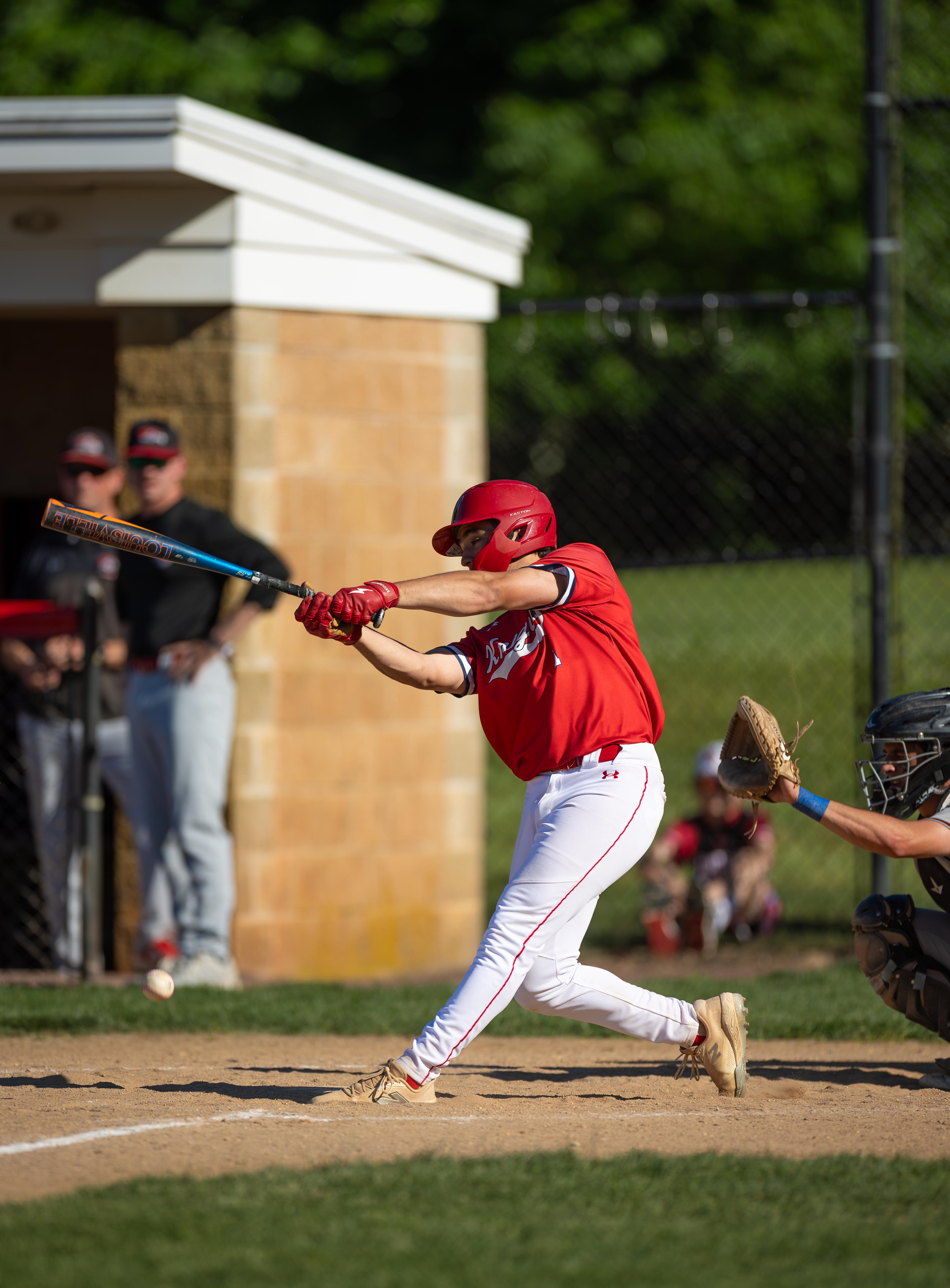 A baseball player in a red uniform swings at a pitch in the foreground, with a dugout and spectators visible in the blurred background.