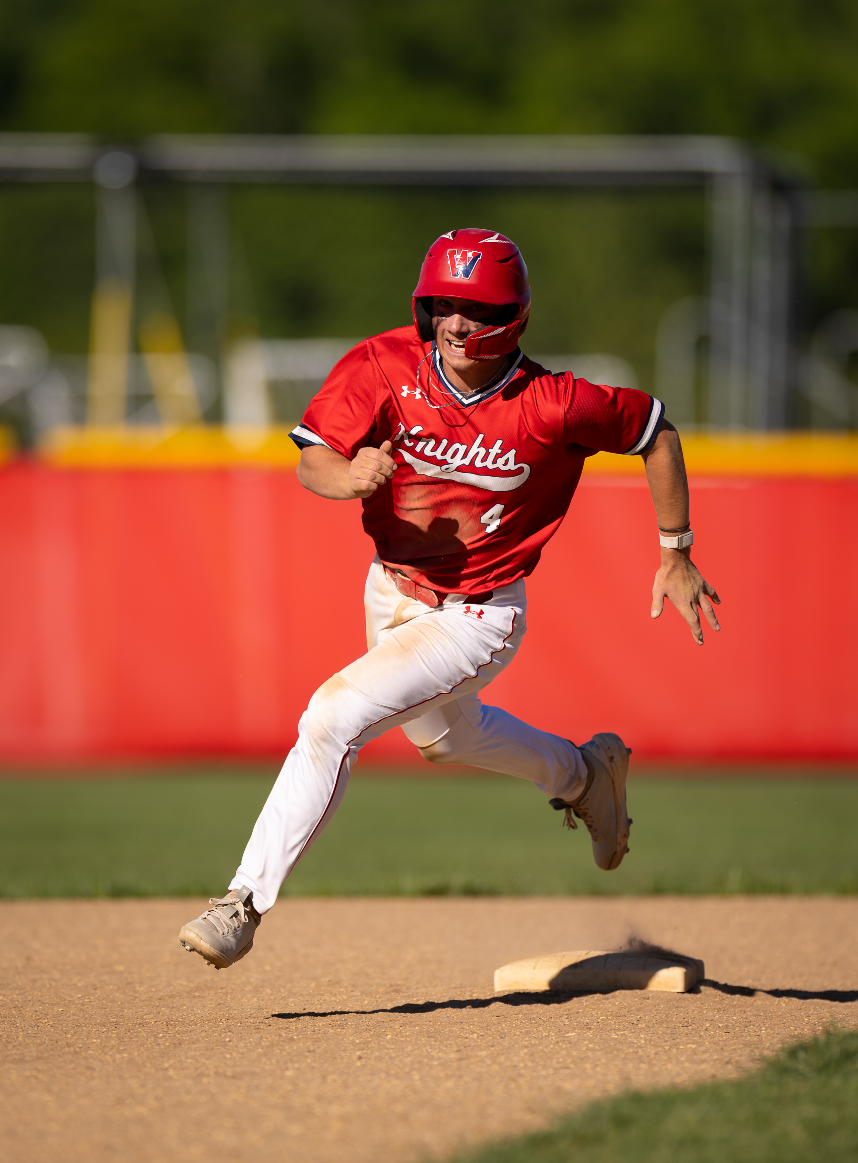 A baseball player in a red uniform runs past a base on a dirt infield with a blurred red outfield fence and green trees in the background.