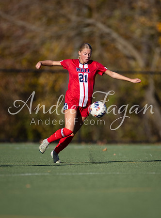 A female soccer player in a red uniform kicks a ball on a green field with trees in the blurred background.