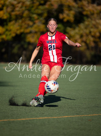 A female soccer player in a red and white uniform kicks a ball on a green field, with autumn trees in the background.