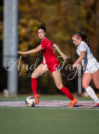 A female soccer player in a red jersey dribbles the ball on a green field with another player in a white jersey behind her, while autumn trees form a blurred background.