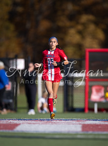 A young woman in a red soccer uniform runs on a field with blurred spectators and trees in the background.