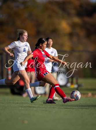 A soccer player in red kicks the ball on a field with other players and trees in the background.