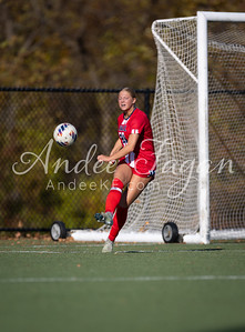 A female soccer player in a red uniform kicks a ball near a goal on a grassy field with trees in the background.