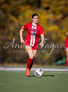 A young woman in a red soccer uniform dribbles a ball on a green field with blurred trees in the background.
