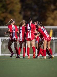 A group of female soccer players in red and white uniforms huddle together on a green field with a blurred background of trees and a fence.