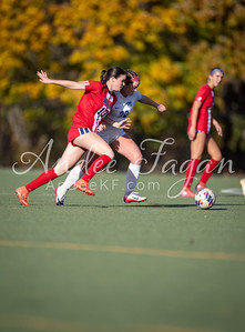 Two female soccer players in red and gray uniforms chase a soccer ball on a green field with a blurred background of yellow and green foliage.