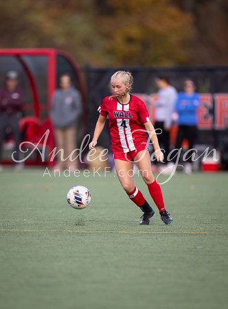 A young woman in a red soccer uniform dribbles a ball on a green field, with blurred figures and a red bench in the background.