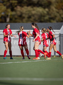 Five female soccer players in red and white uniforms are on a green field with a building and trees in the background.